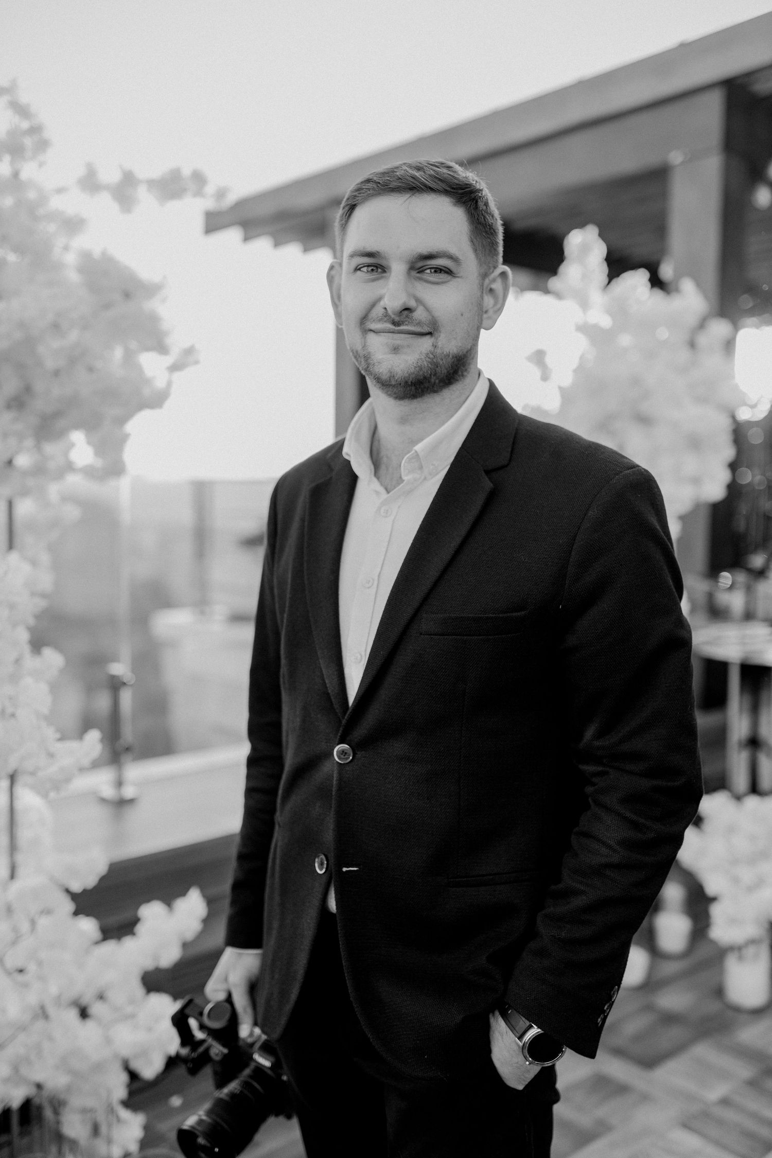 Black and white portrait of a smiling man in a suit holding a camera at an outdoor event.