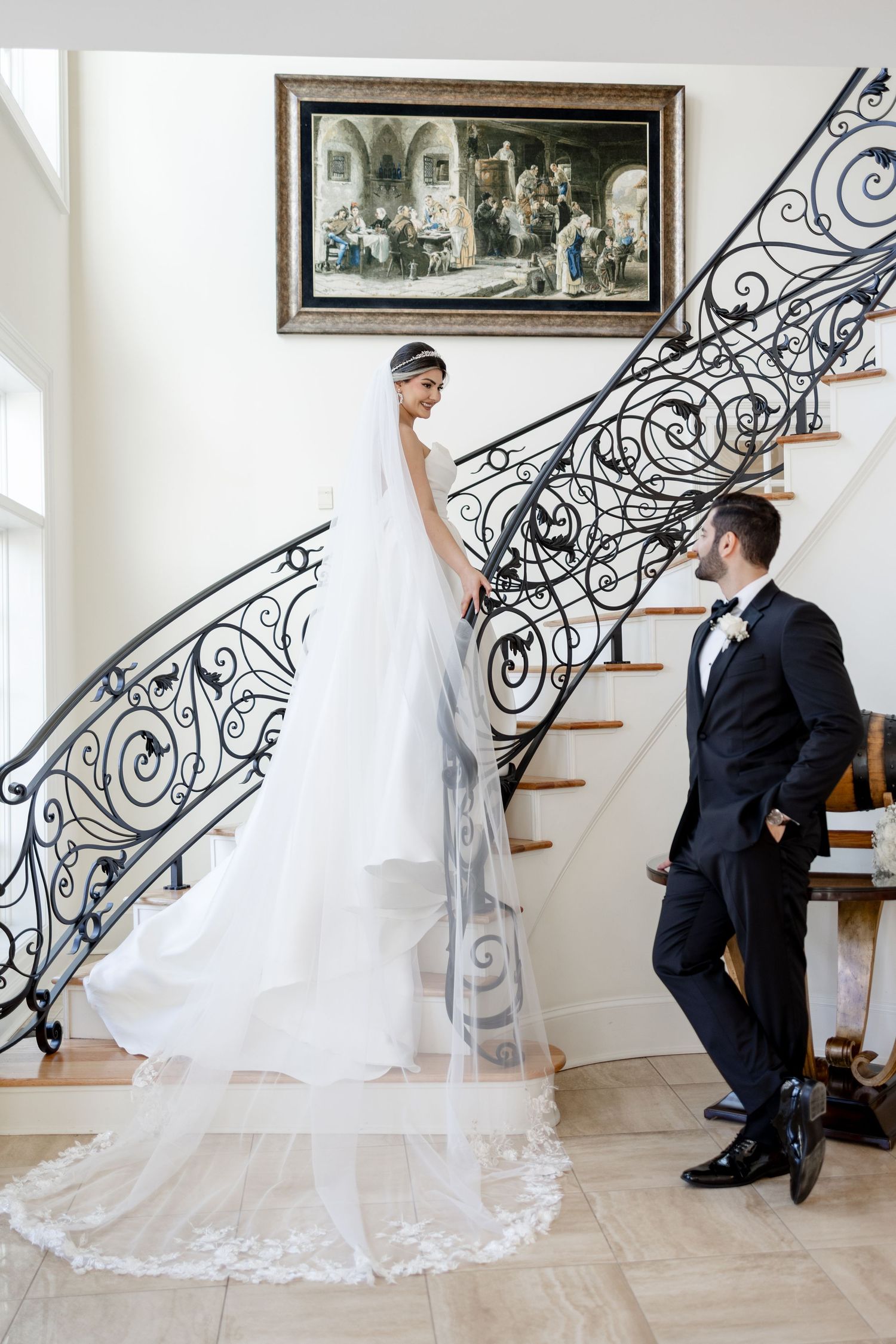 Bride in white gown with cathedral veil stands on elegant staircase as groom in black tuxedo gazes up admiringly.