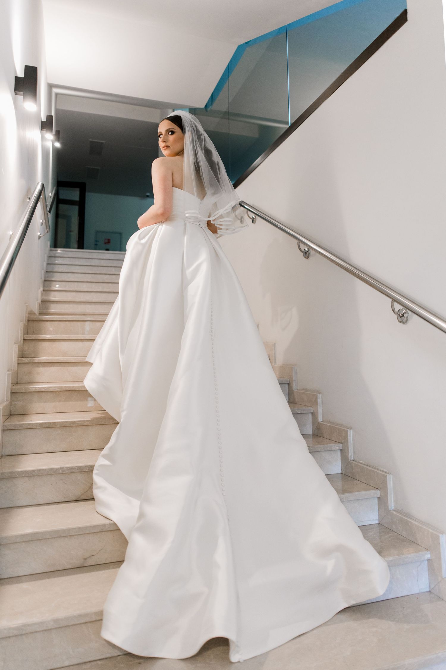 Elegant bride in a white strapless ball gown with veil ascending marble stairs, viewed from behind.