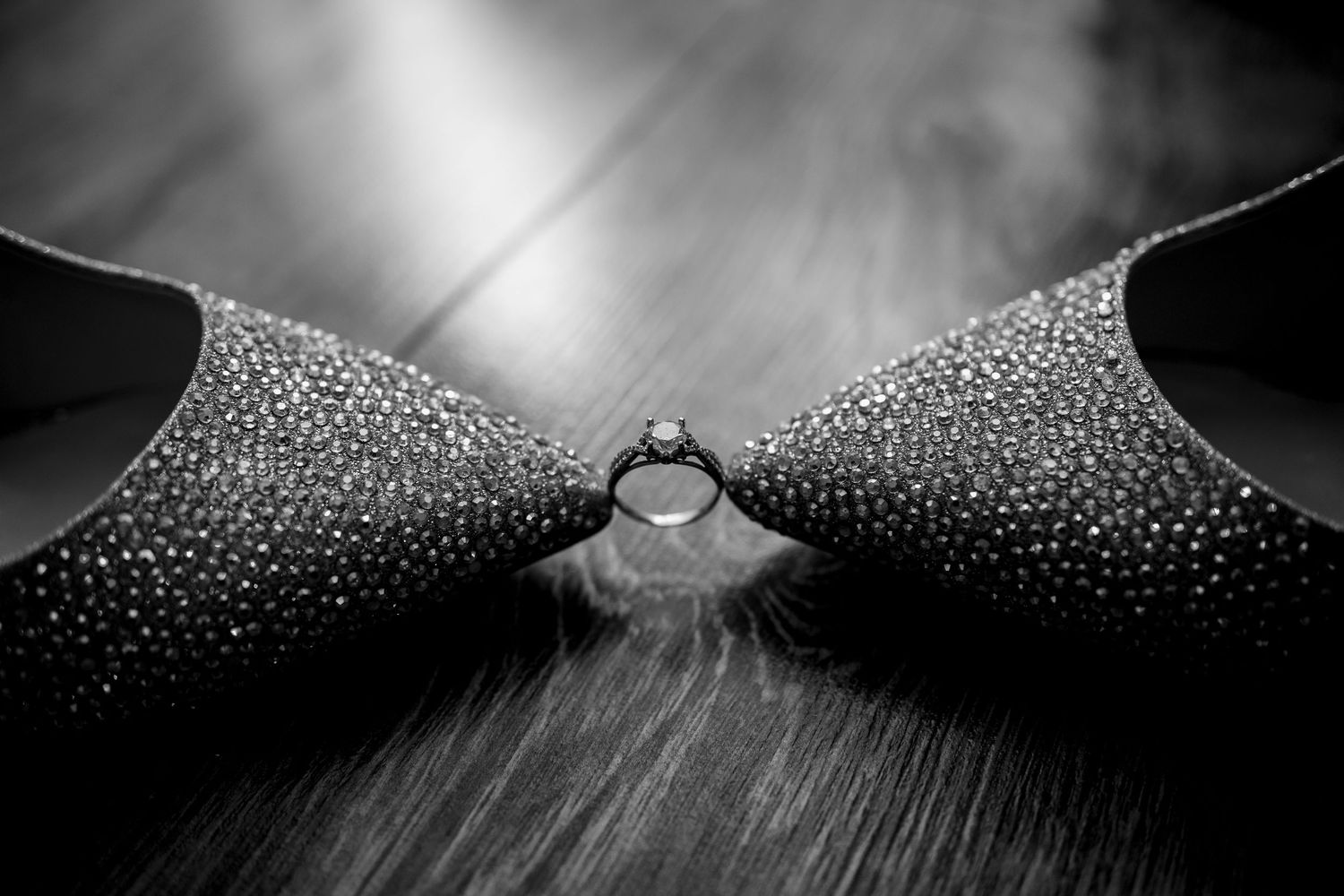 Black and white photo of a diamond engagement ring placed between two rhinestone-studded high heel shoes on a wooden floor.