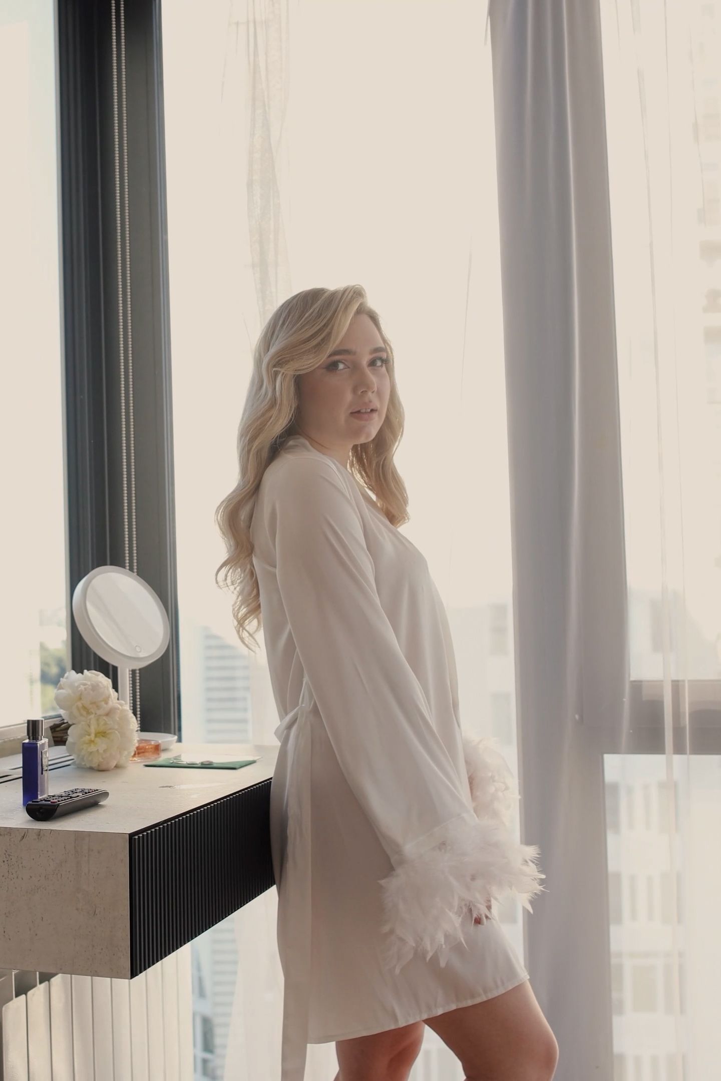 Blonde woman in white feather-trimmed robe standing by vanity mirror near large window in bright room.