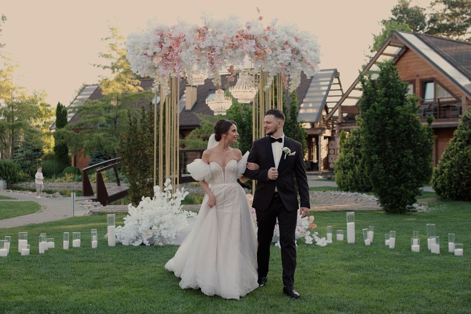 Bride and groom stand together outdoors at elegant wedding ceremony with floral arch and candles on green lawn.