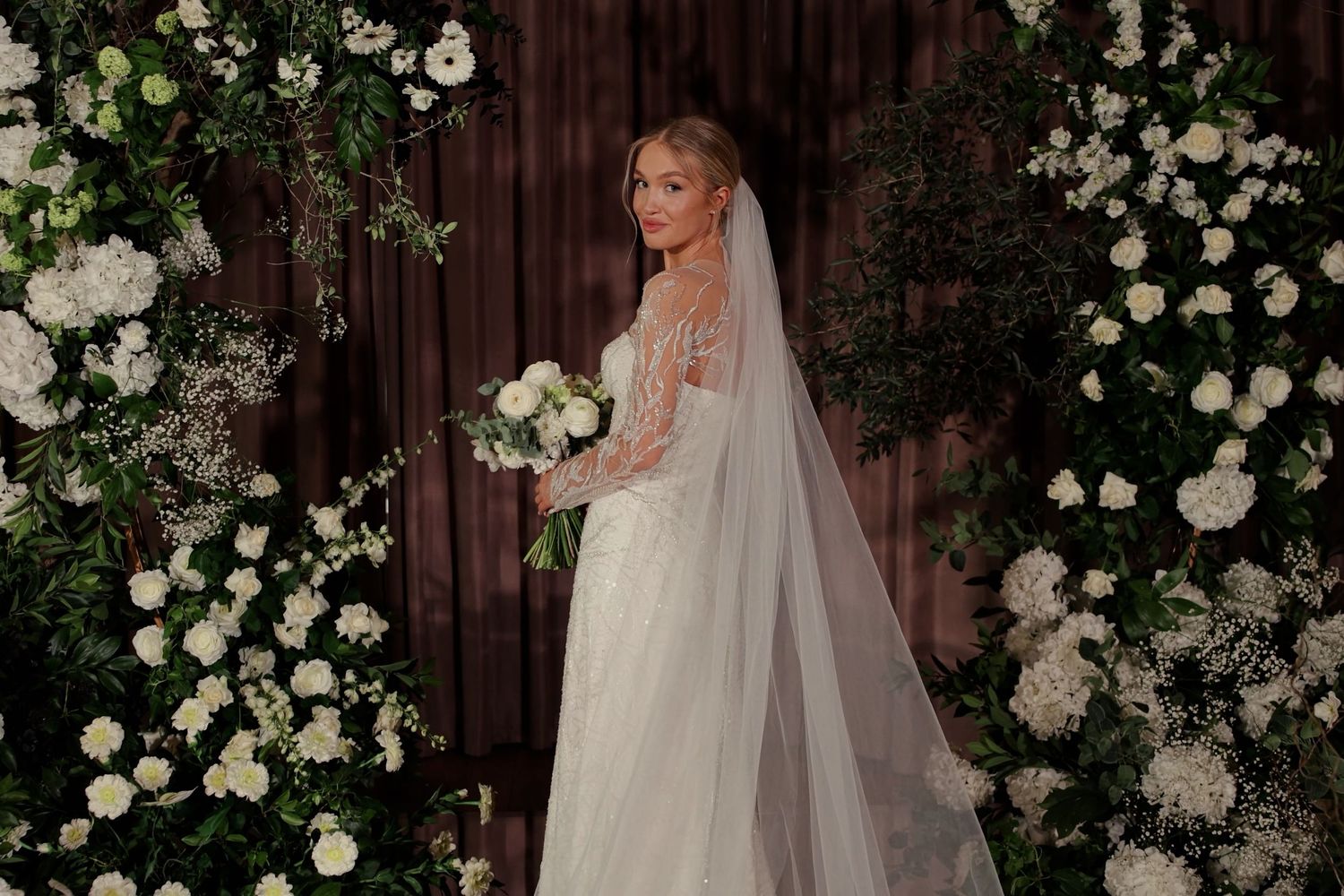 Elegant bride in a beaded long-sleeve wedding gown and veil poses before a lush white floral arch backdrop.