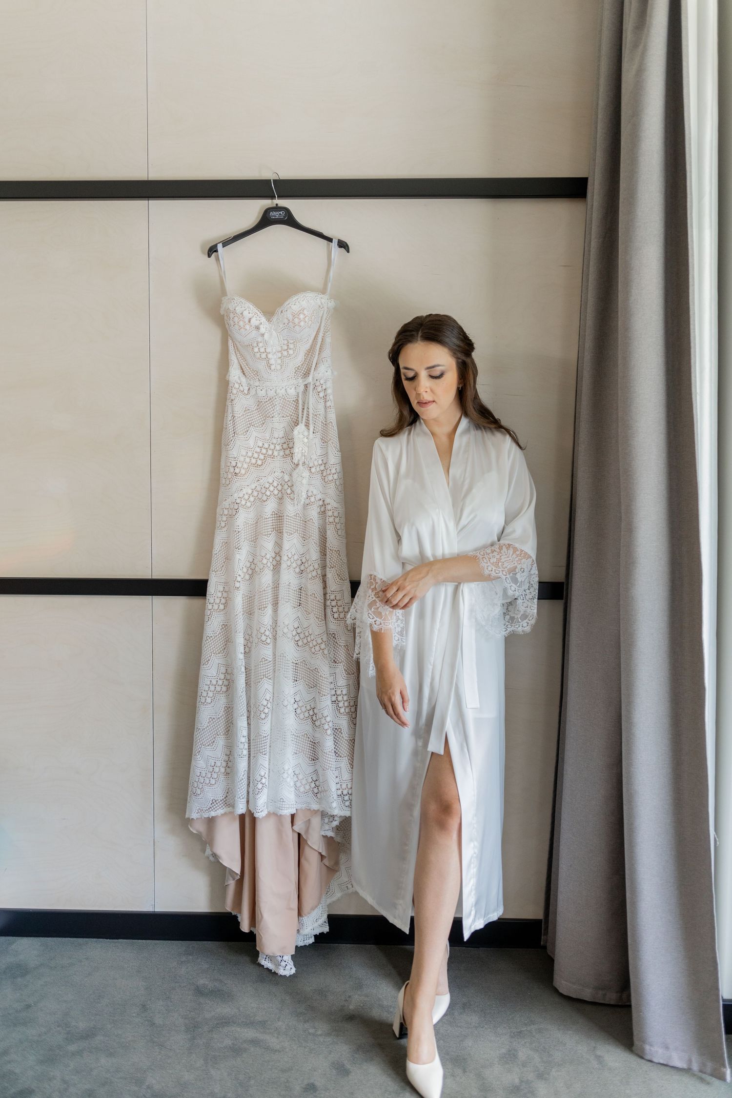 A white lace wedding dress hangs on display next to someone in a white robe in a dressing room.