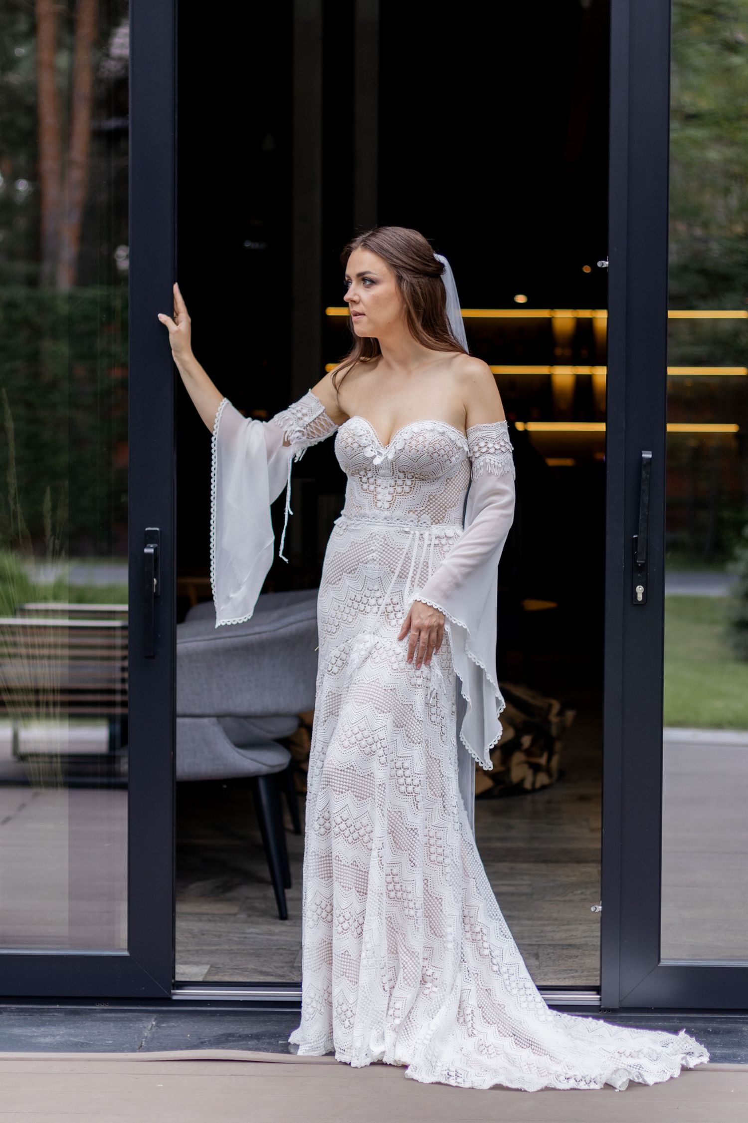 Elegant bride poses in flowing lace wedding gown against modern glass doorway.