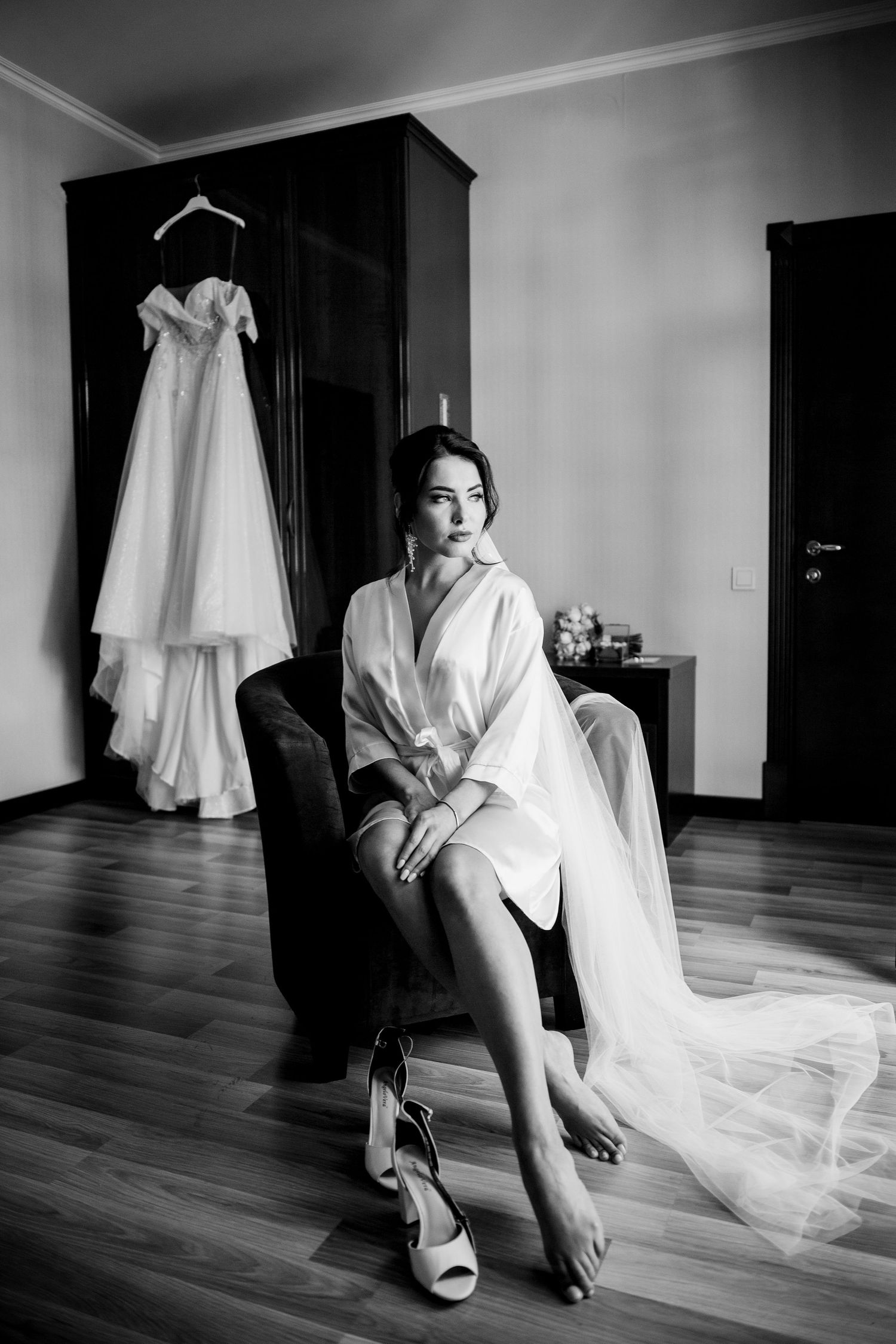 Black and white photo of a bride in a white robe sitting in a chair with her wedding dress hanging in the background.