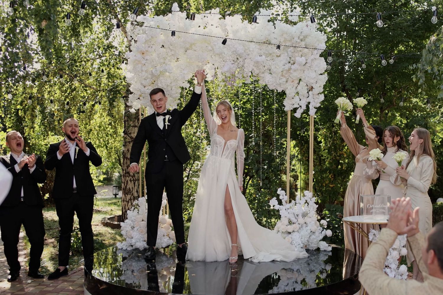 Bride and groom celebrate under white floral arch at outdoor wedding ceremony with bridesmaids and groomsmen cheering.