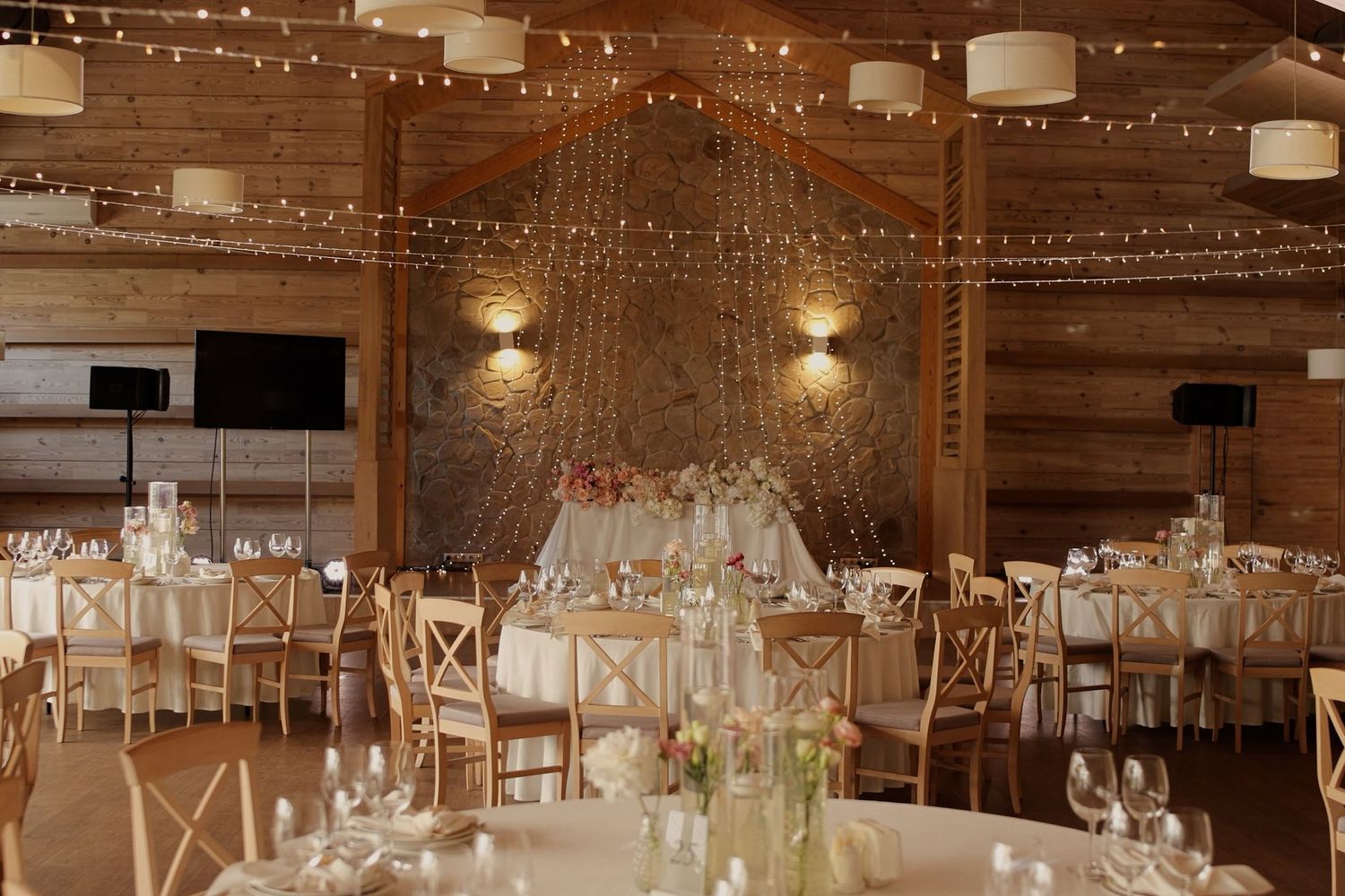 Rustic barn wedding reception with string lights, wooden chairs, round tables, and elegant floral centerpieces.