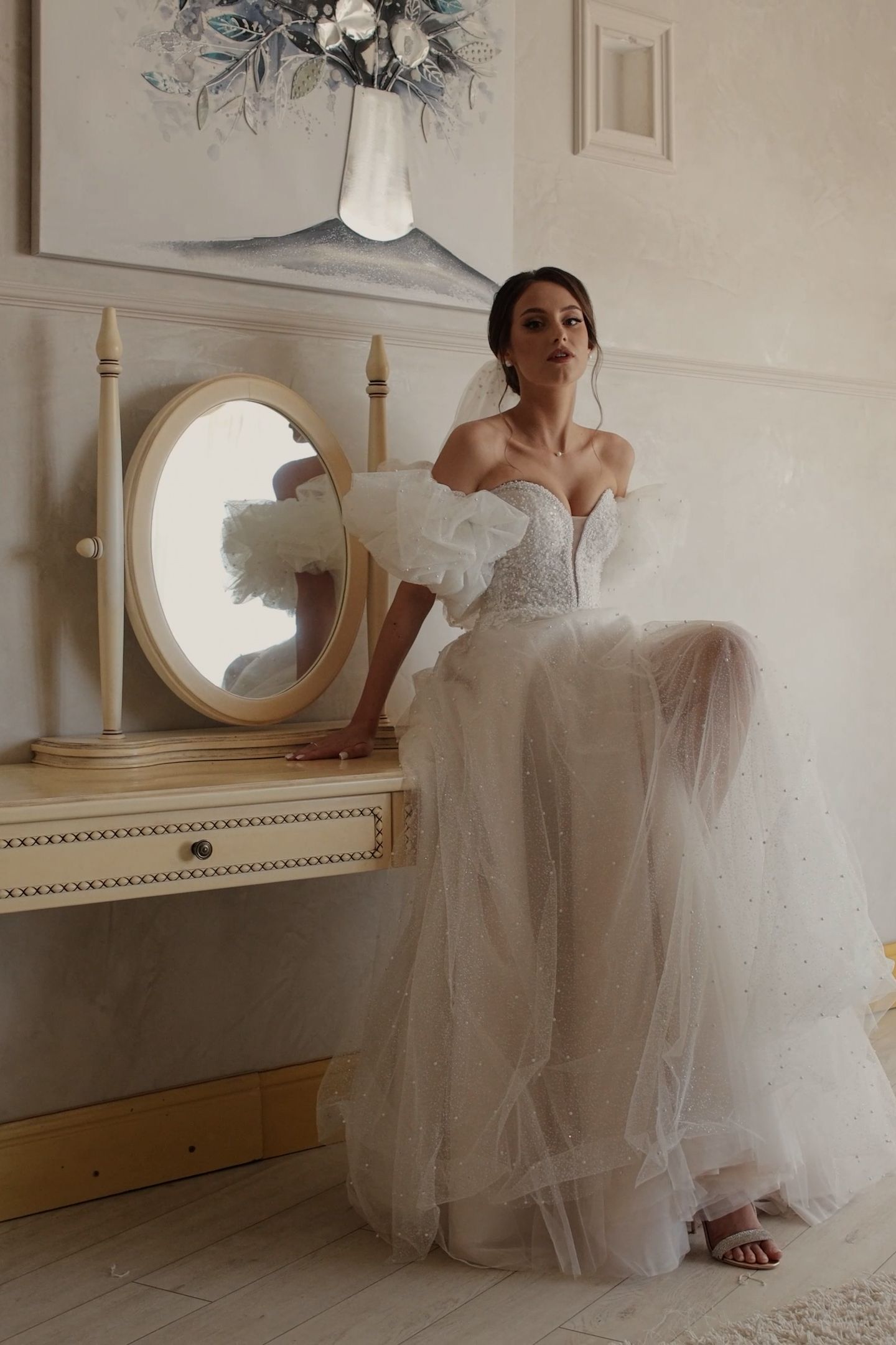 Bride in off-shoulder white tulle wedding gown sits at vintage vanity mirror in elegant room.