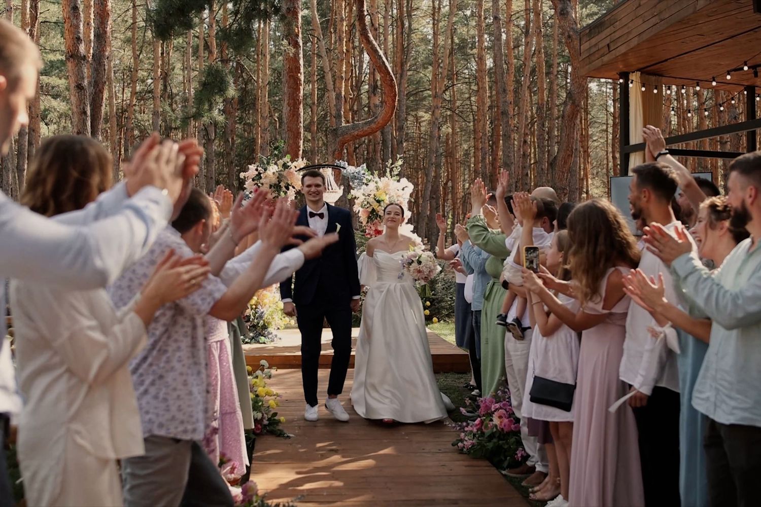 Bride and groom walk through applauding guests at outdoor forest wedding ceremony with floral decorations.