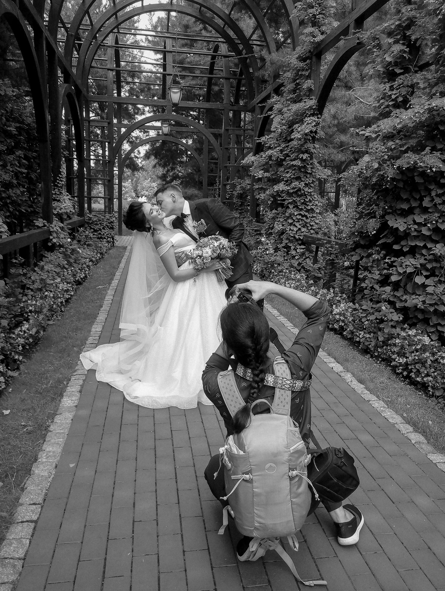 Black and white photo of a wedding couple kissing under a garden arbor while a photographer crouches to capture the moment.