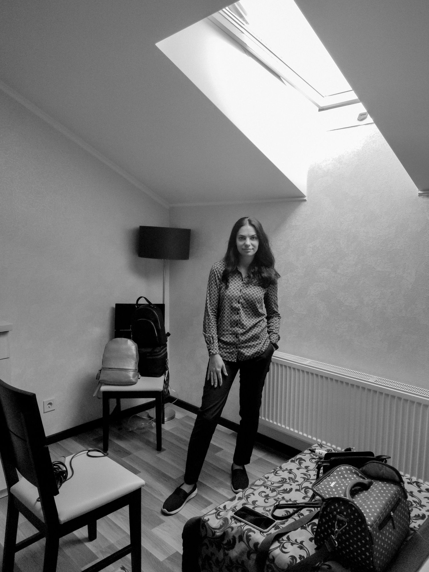 Black and white photo of a woman standing in a cozy attic room with skylight, chairs, and luggage on the floor.
