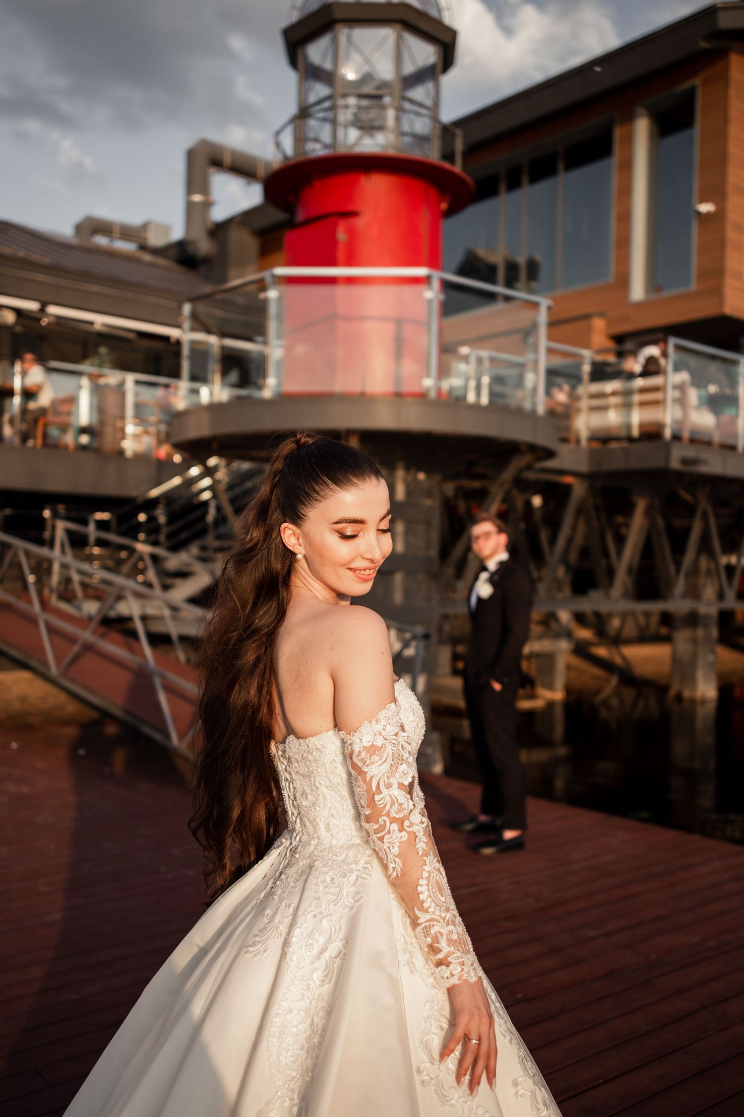 Bride in lace off-shoulder gown poses near red lighthouse at waterfront venue with groom in background.