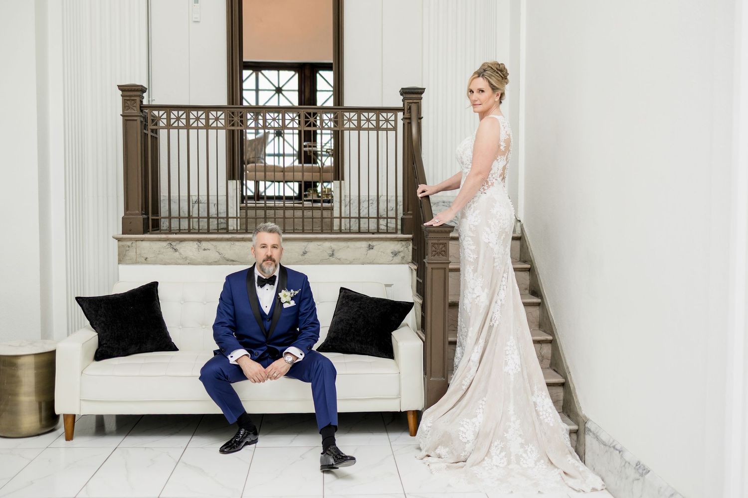 Bride in lace gown stands on staircase behind groom in navy suit seated on white sofa in elegant venue.