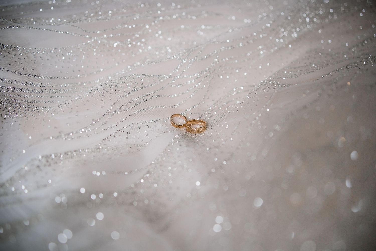 Two gold wedding rings resting on a sparkling white bridal veil with glittering bokeh details.