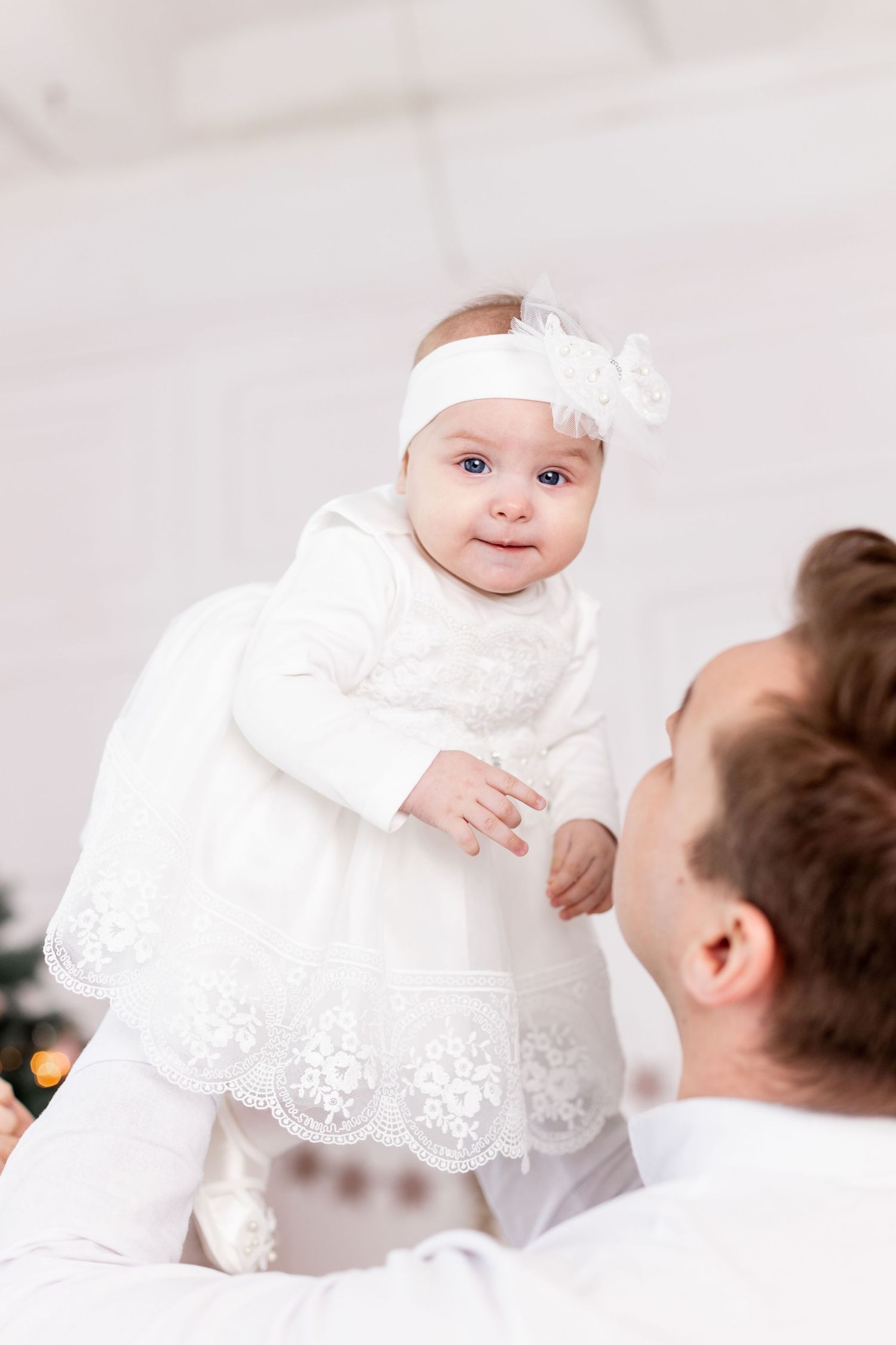Baby in white lace christening gown and headband being lifted up against light background.