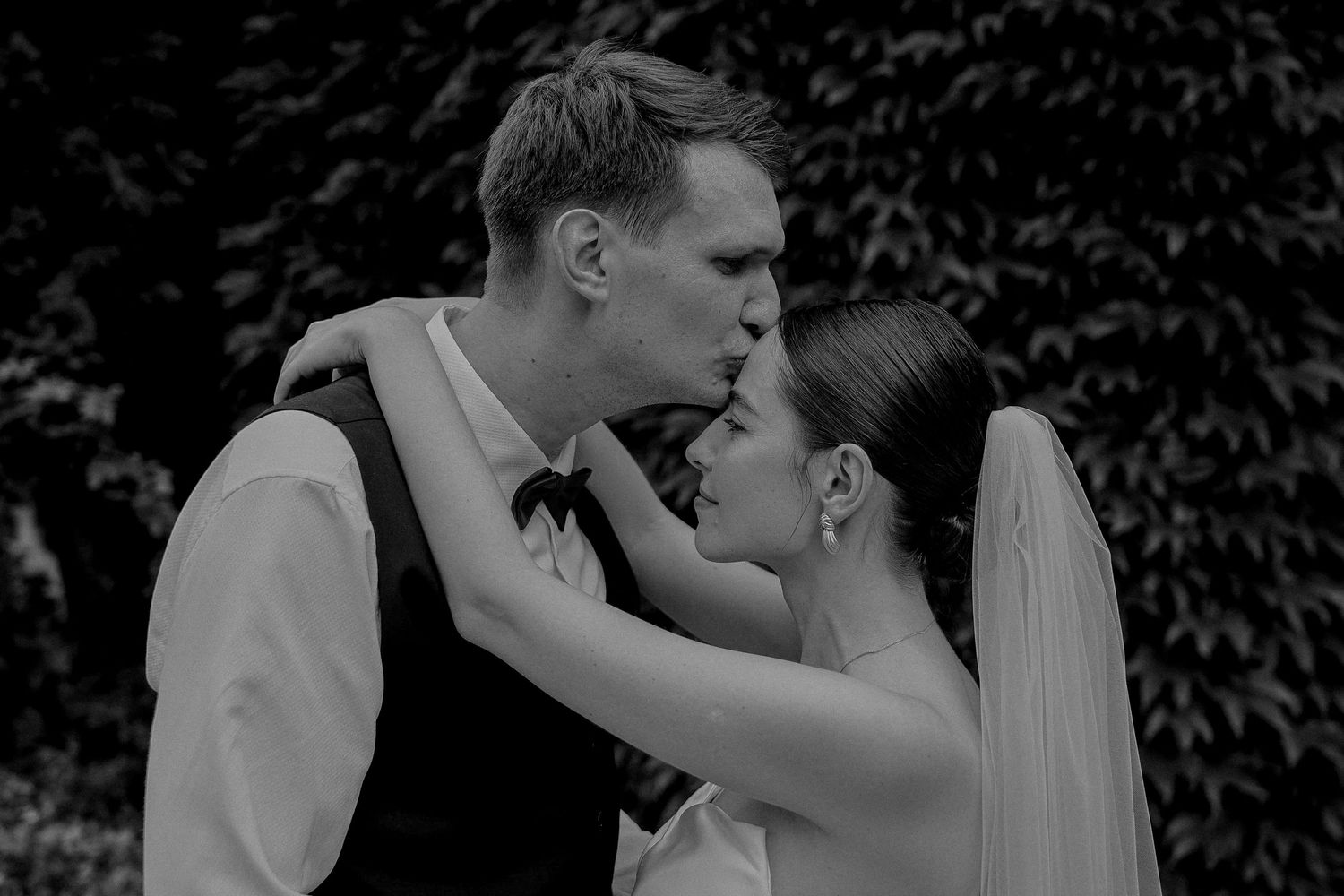 Black and white photo of a bride and groom sharing a romantic kiss outdoors on their wedding day.