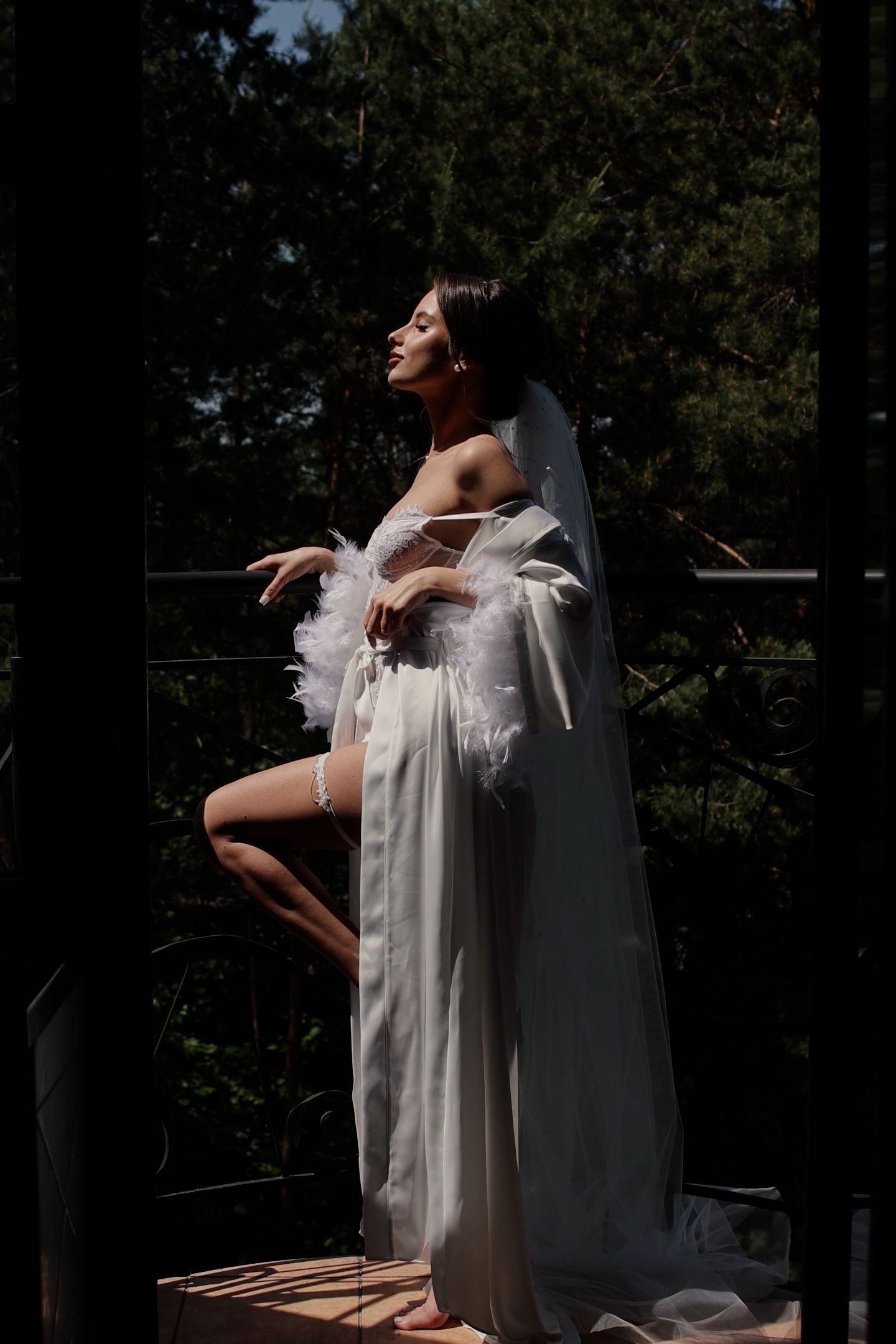 Elegant bride in white sheer robe and veil poses gracefully on outdoor balcony surrounded by lush green trees.