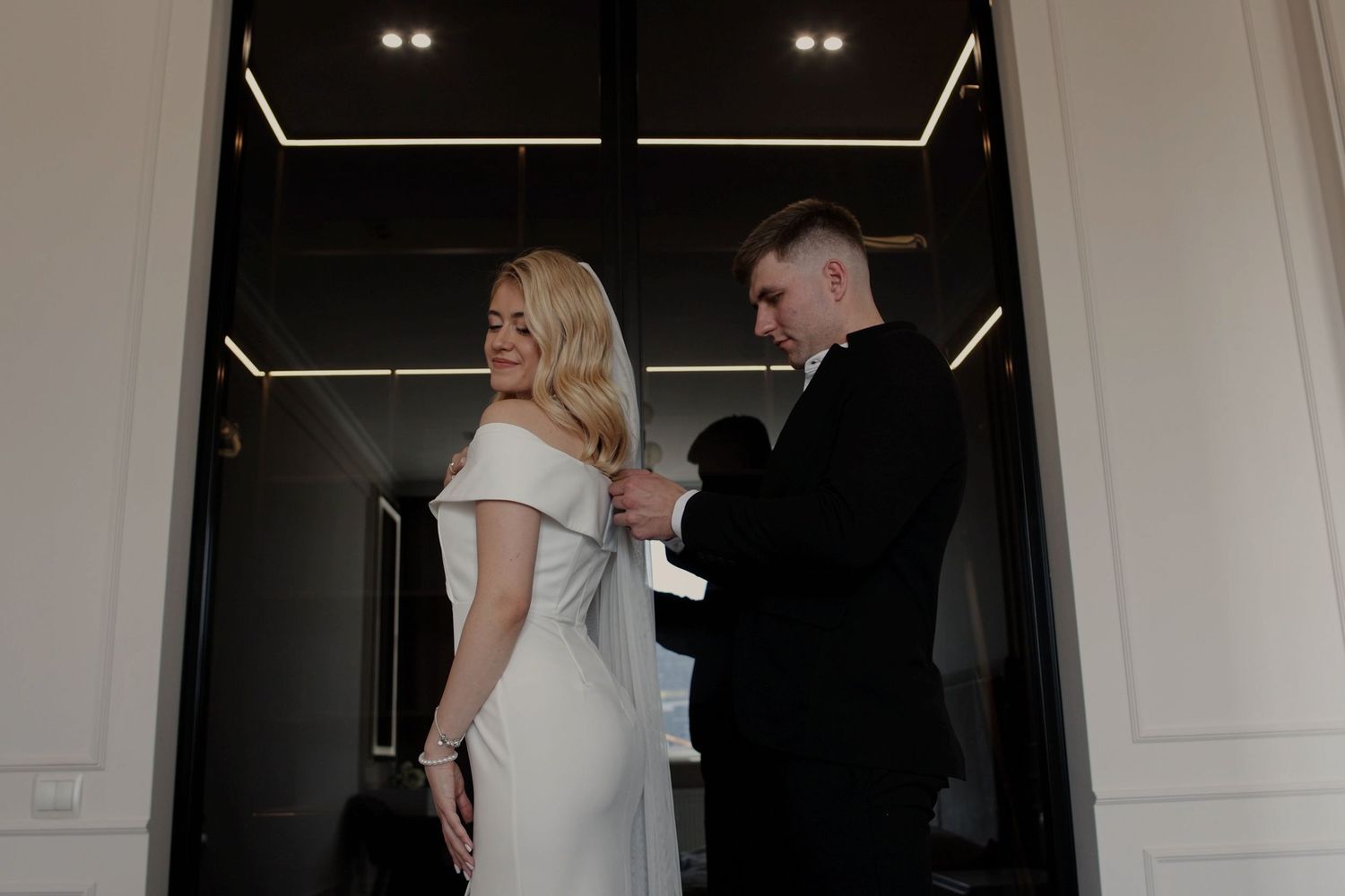 Groom in black suit fastening the back of bride's white off-shoulder wedding dress in elegant white room.