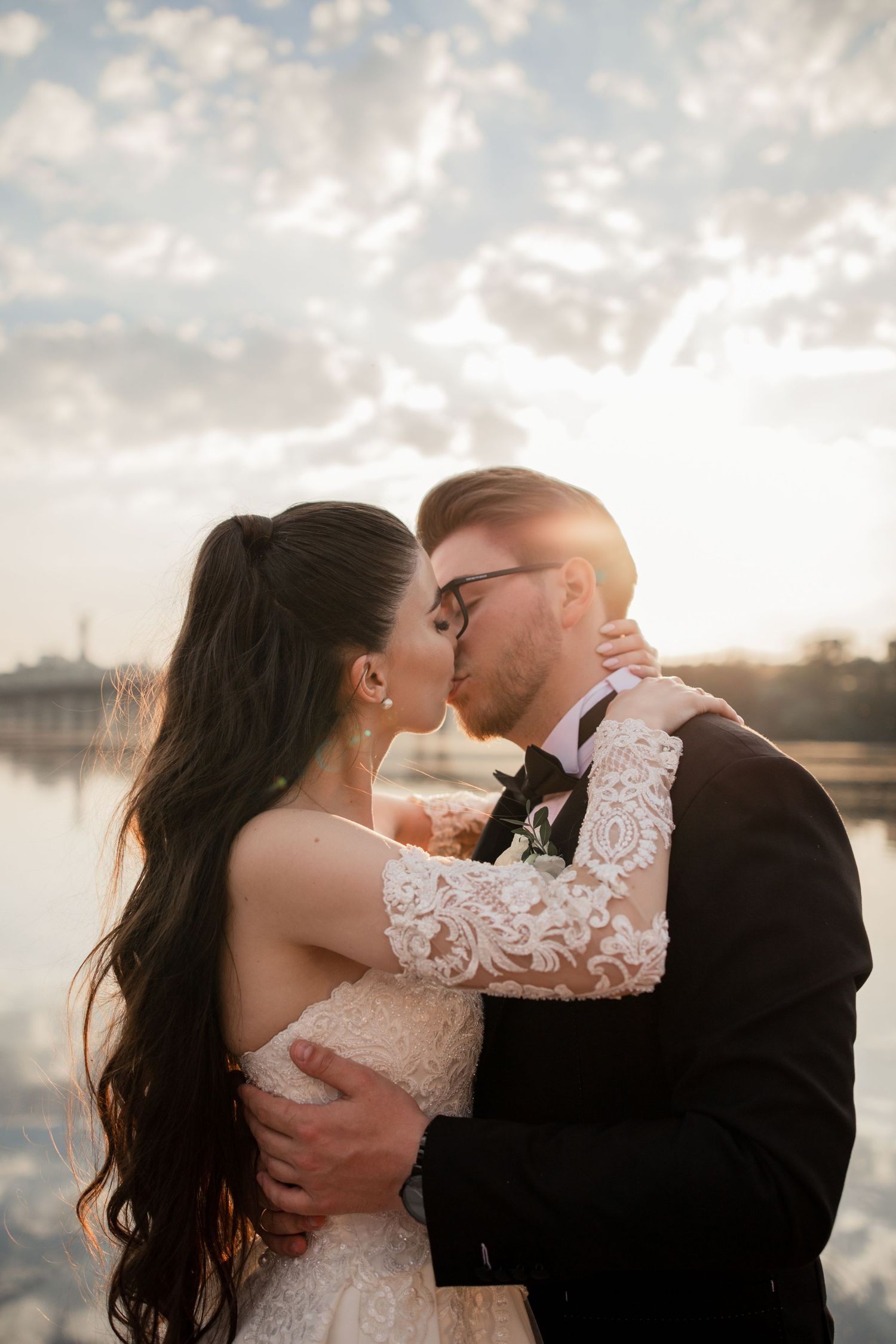 A bride and groom share a romantic kiss at sunset by a waterfront, she in lace gown, he in black tuxedo.
