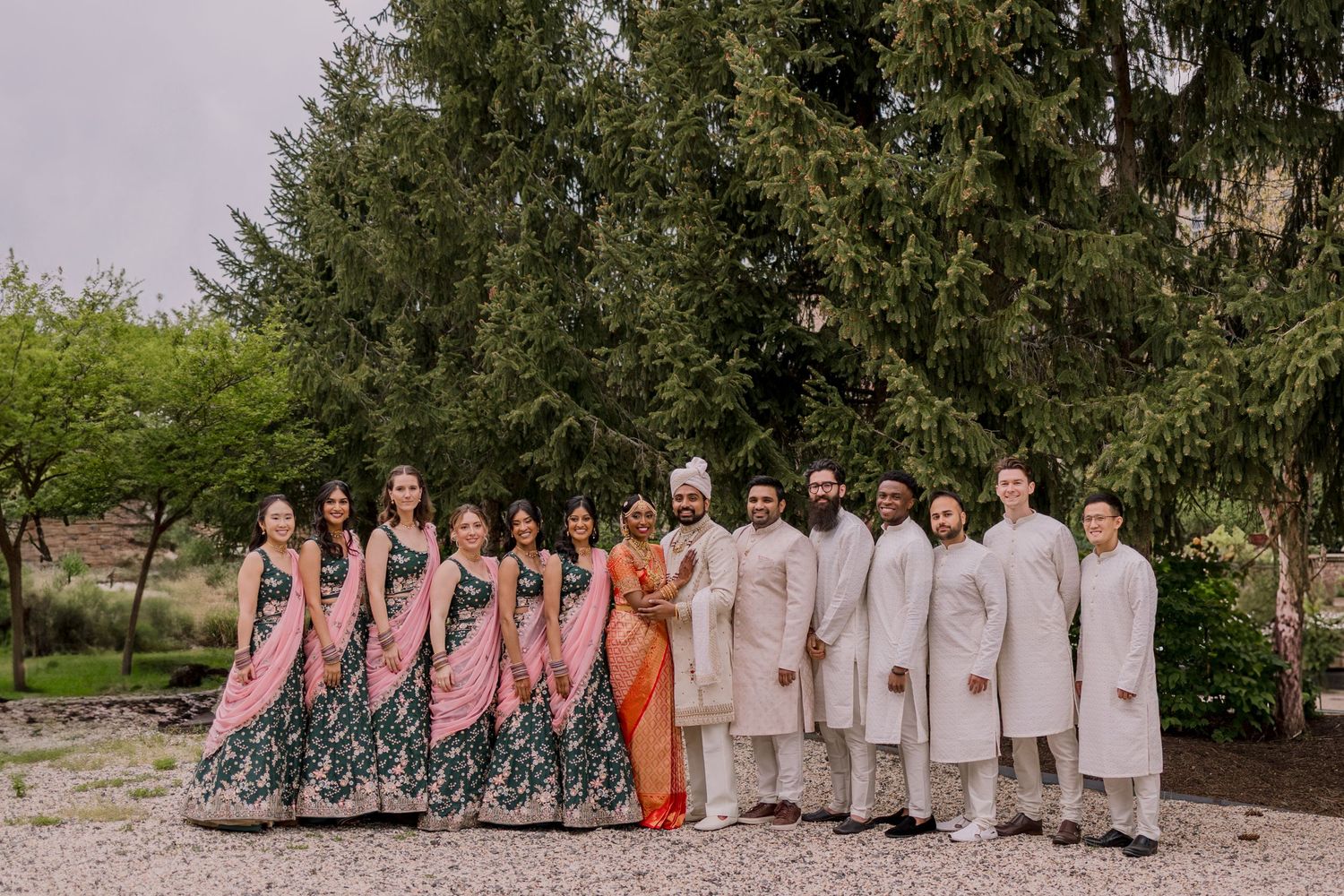 Indian wedding party posing outdoors in traditional attire, bridesmaids in pink and green lehengas, groomsmen in cream sherwanis.