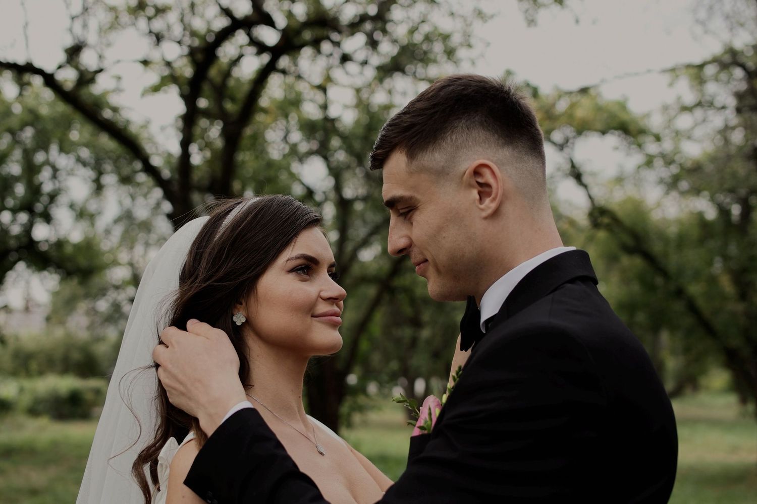 A bride and groom share a romantic outdoor moment, with the bride in a veil and groom in a black suit.