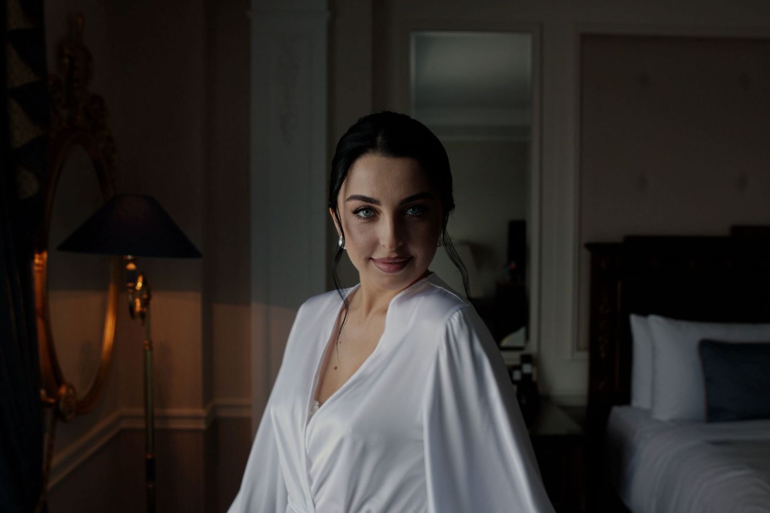 A woman in a white satin robe smiles softly in a dimly lit elegant hotel room.