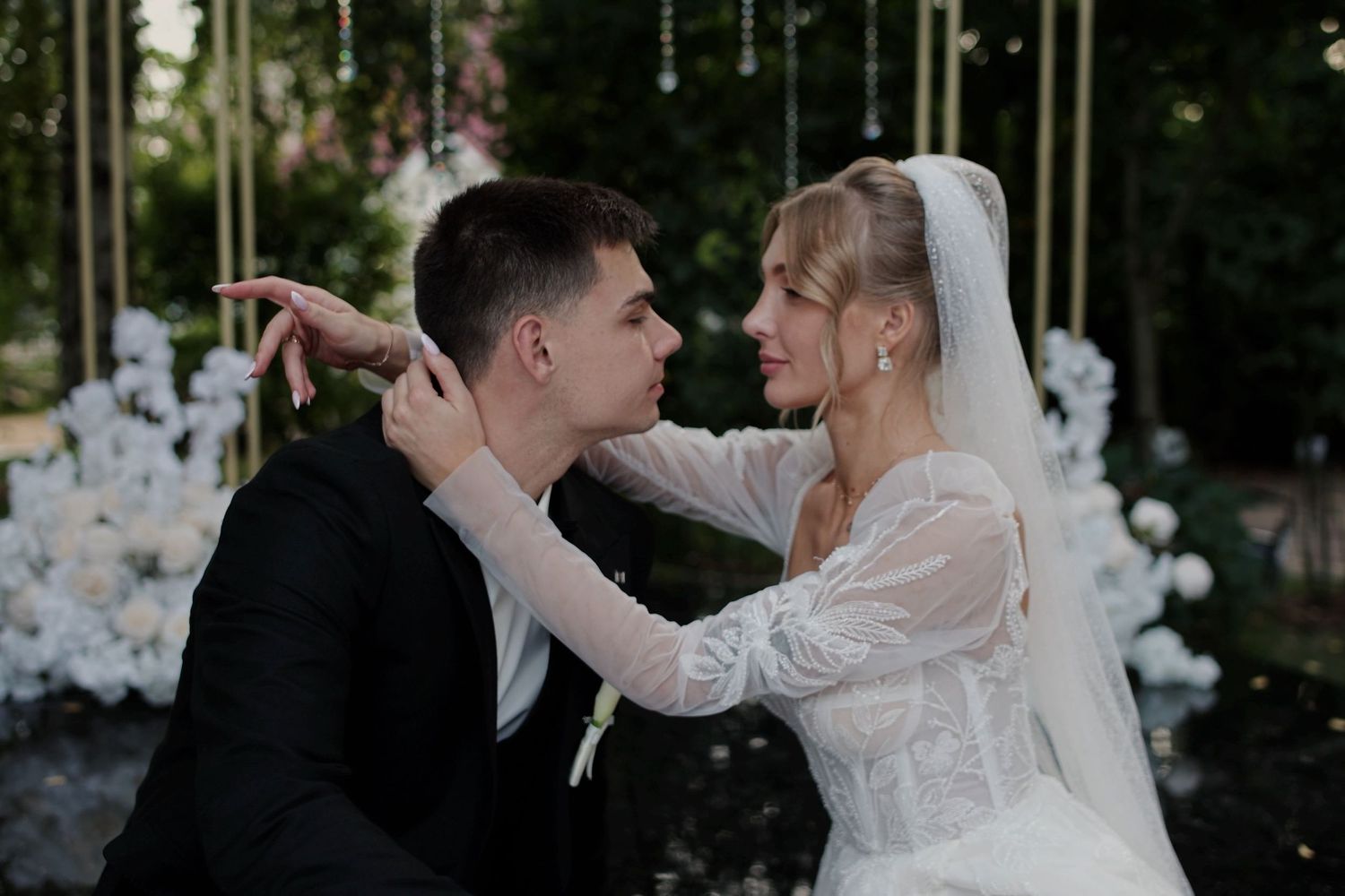 Bride in white lace gown and veil tenderly touches grooms face during romantic outdoor wedding ceremony.