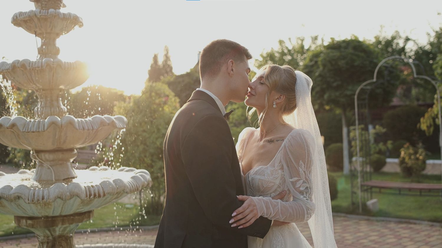 Bride and groom share a romantic kiss near an ornate stone fountain in a sunlit garden on their wedding day.