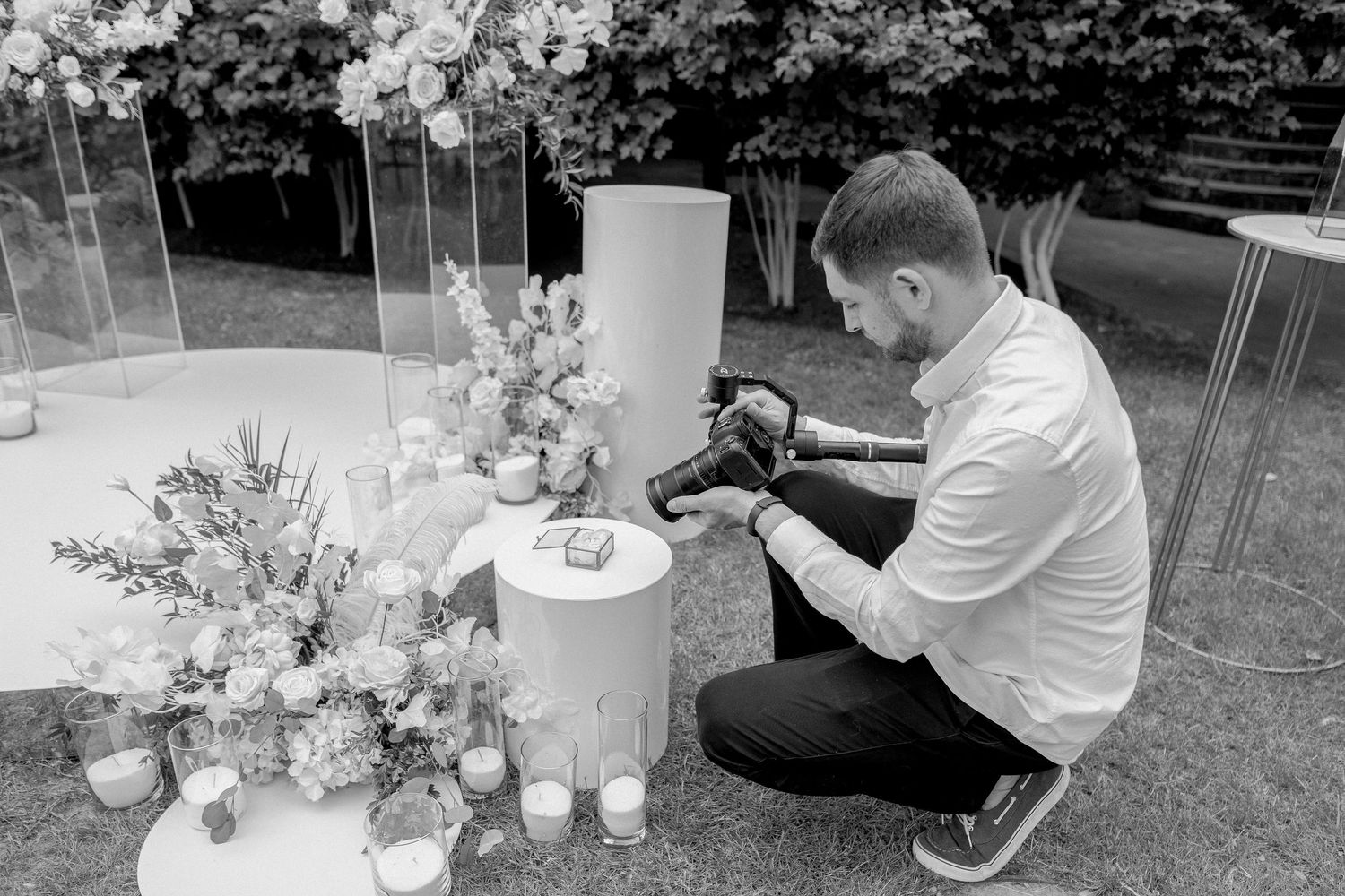 Black and white photo of a male photographer crouching to photograph wedding decor with flowers and candles.