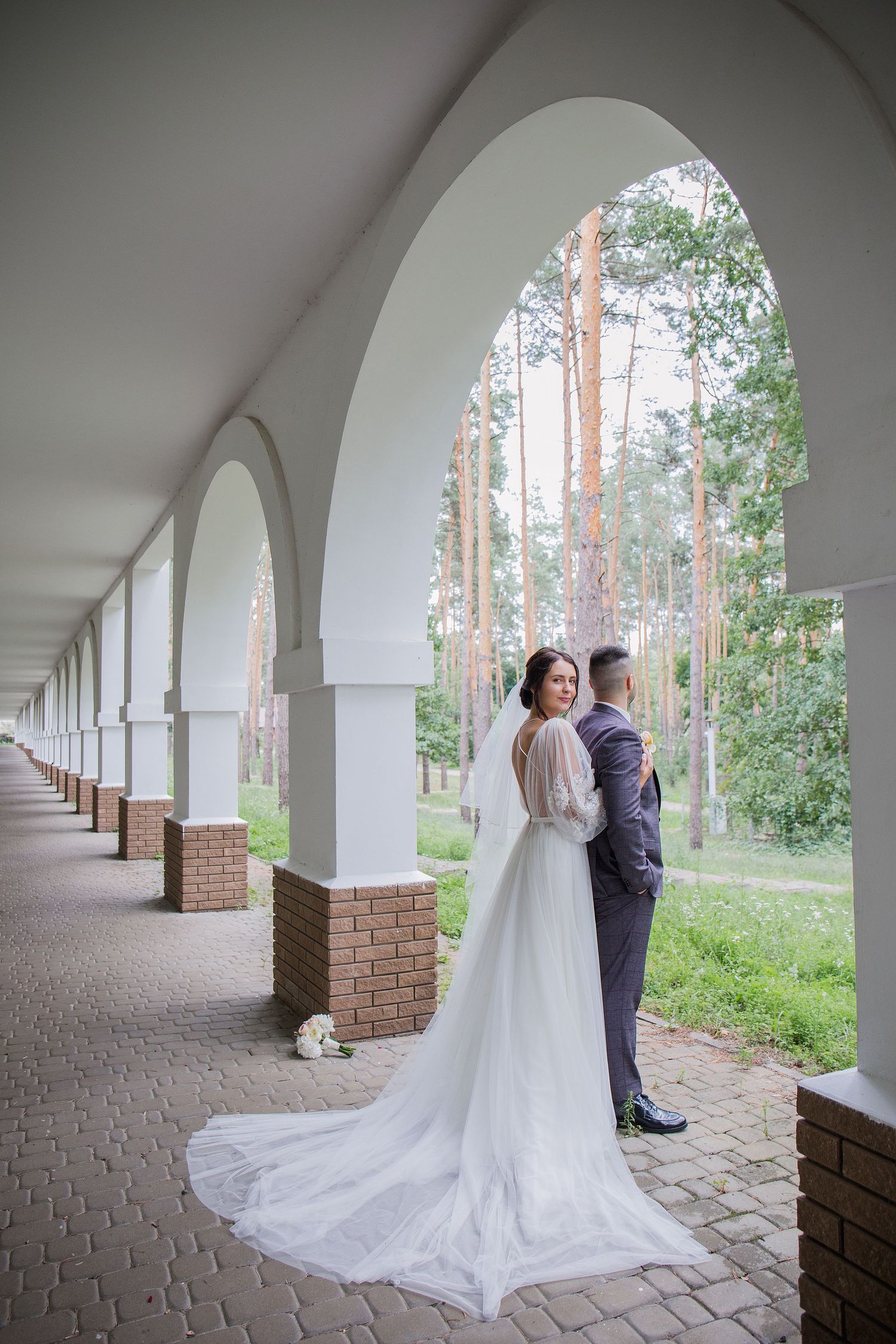 Bride and groom pose under white arched colonnade with brick pillars, green forest backdrop, long wedding dress train.