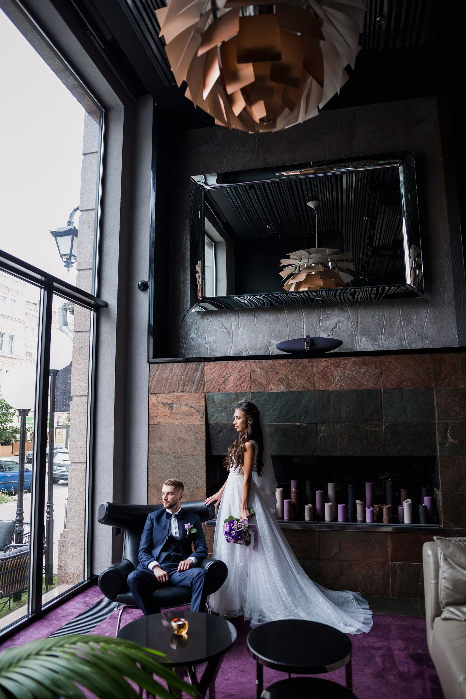 Bride in white gown holds purple bouquet beside groom in blue suit seated in chair inside elegant dark interior.