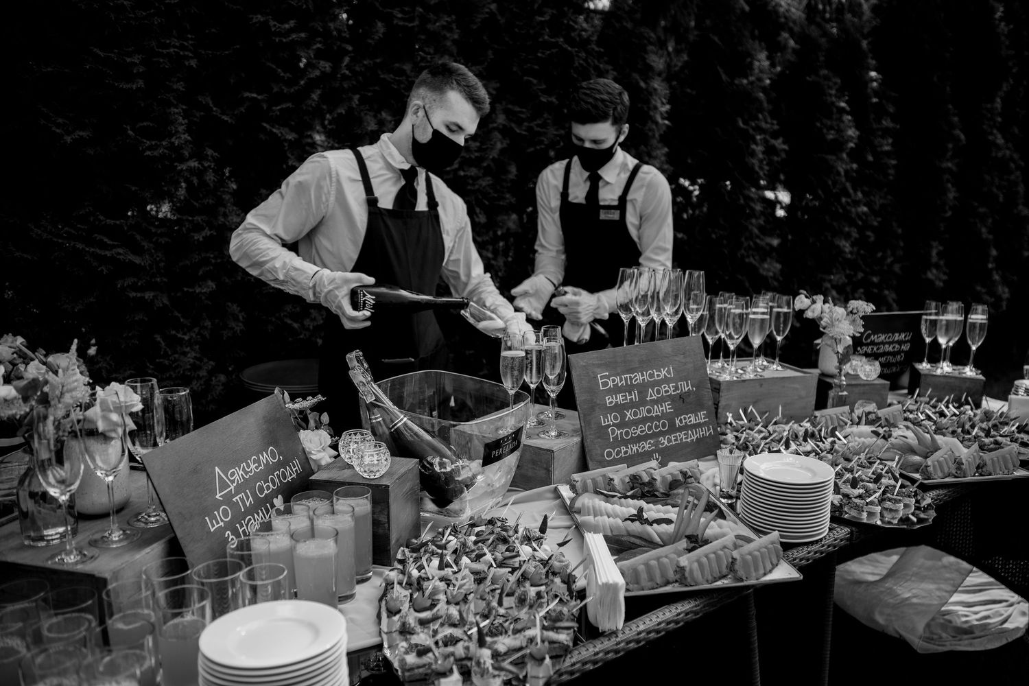 Two masked waiters in aprons arrange food and drinks on an elegant outdoor buffet table in black and white.