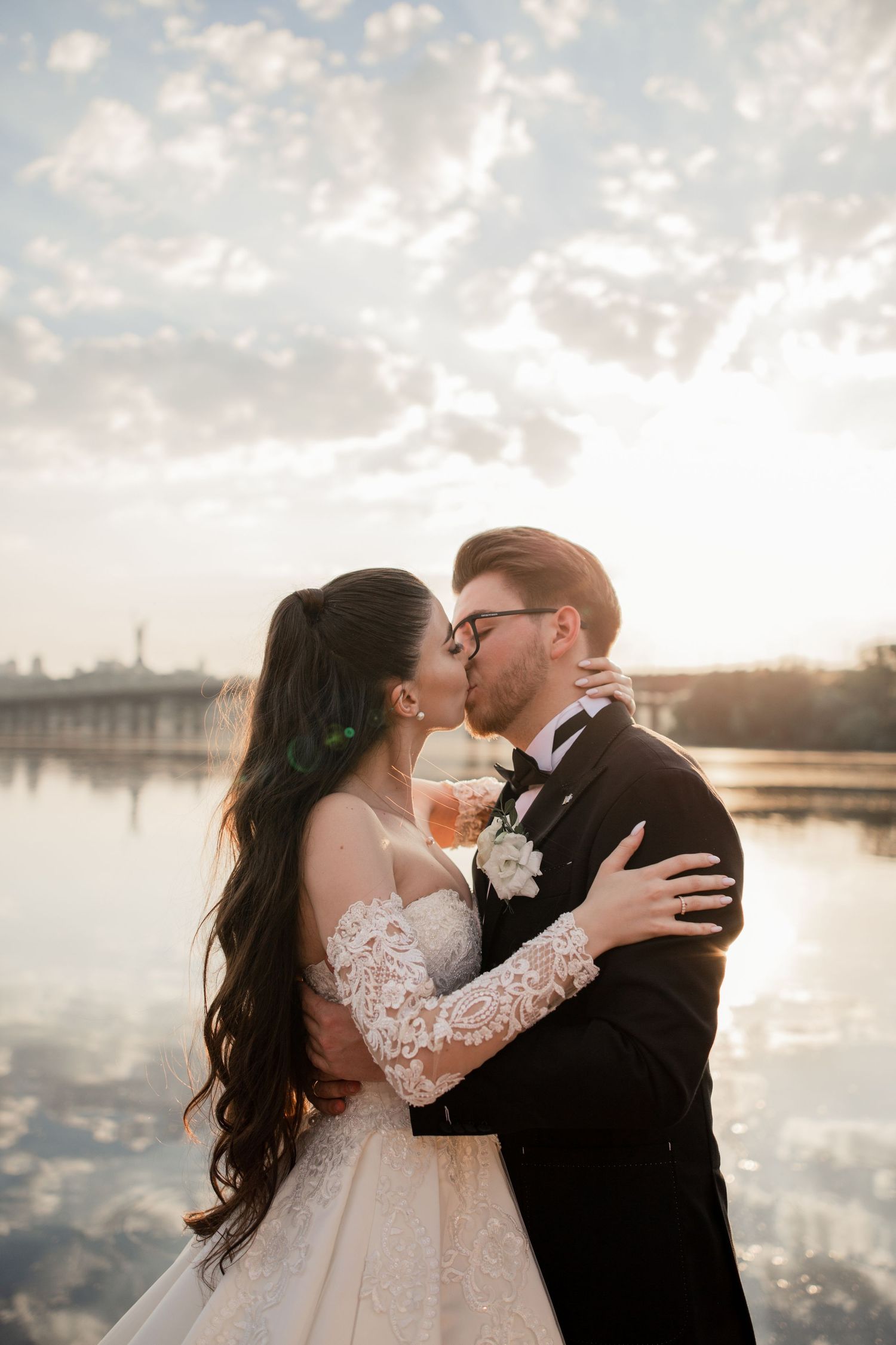 A bride and groom share a romantic kiss at sunset by a serene lake, she in lace gown, he in black tuxedo.