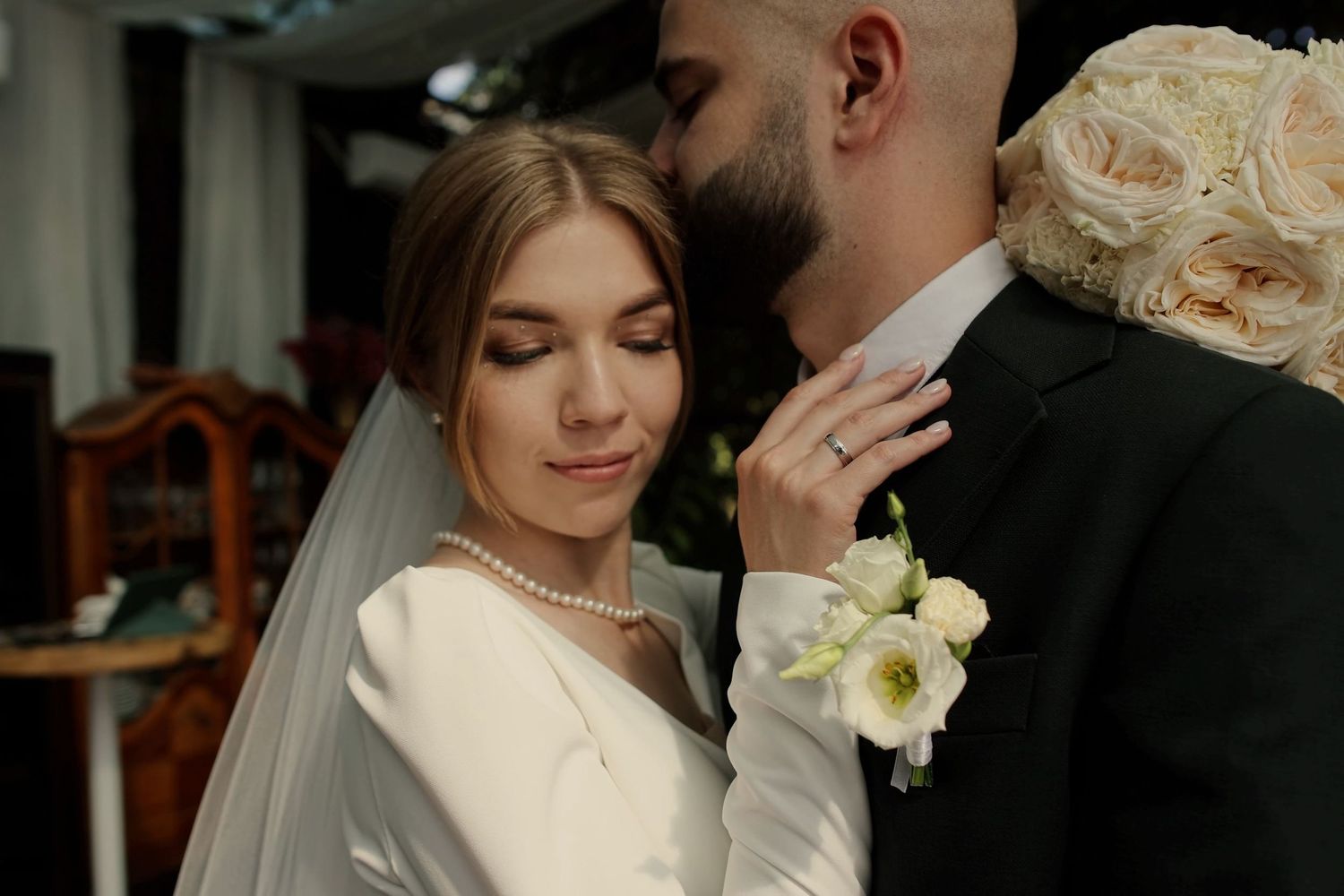 Elegant bride in white gown and veil with pearl necklace embraces groom in black suit during romantic wedding ceremony.
