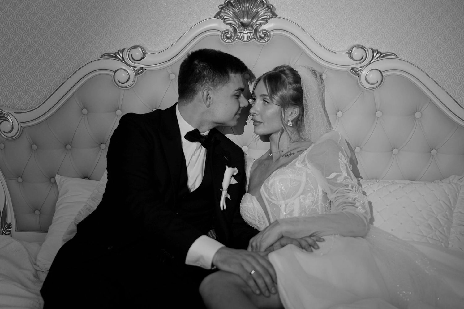 Black and white wedding photo of bride and groom sharing an intimate moment on an elegant tufted bed.