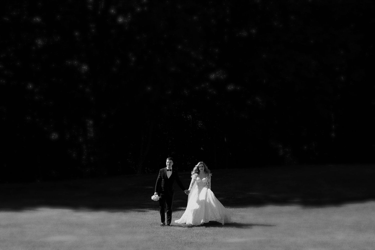 Black and white photo of a bride and groom walking hand in hand across a sunlit lawn with dark trees behind.