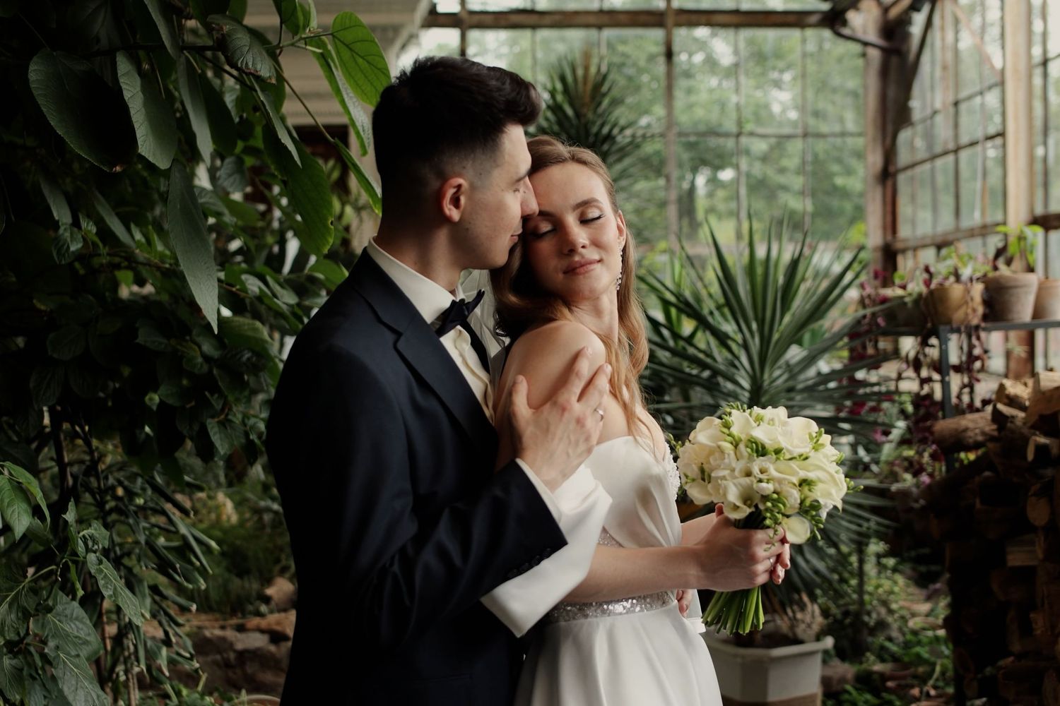 A bride and groom embrace in a lush greenhouse, she holds a white bouquet in a satin gown as he wears a navy suit.