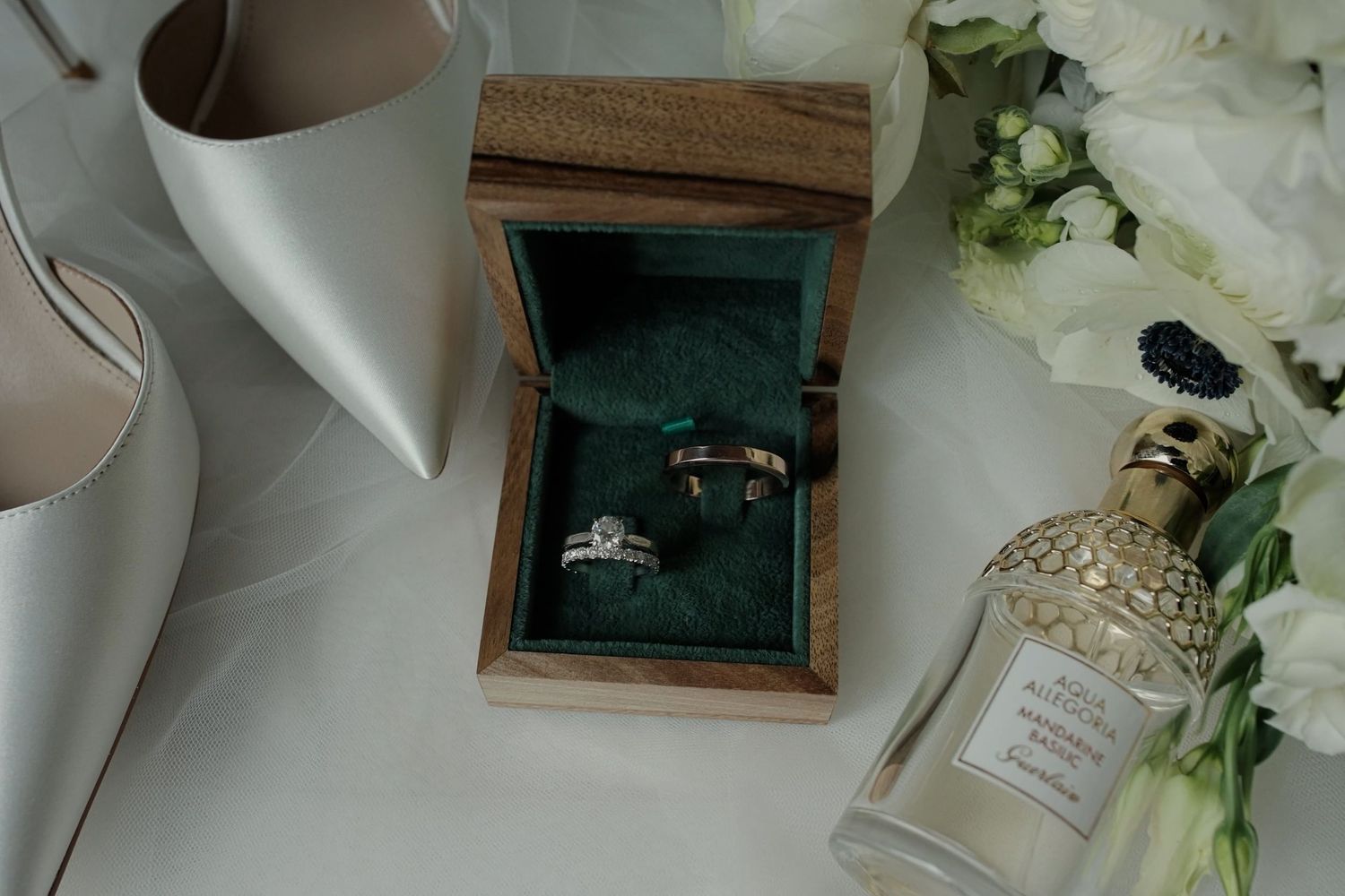 Elegant wedding rings in a green velvet wooden box, beside white heels, florals, and perfume bottle.