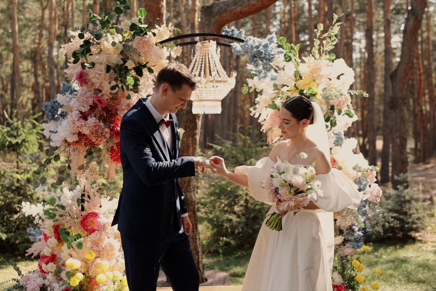 Couple exchanging rings at outdoor wedding ceremony with colorful floral arch and hanging chandelier in forest.