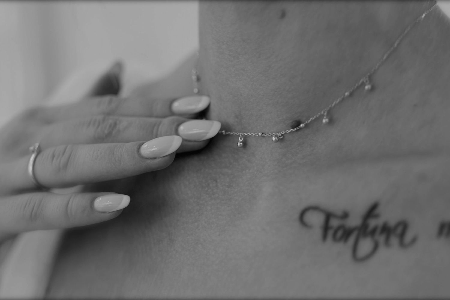 Black and white photo of a woman wearing a delicate chain necklace with a cursive tattoo on her chest.