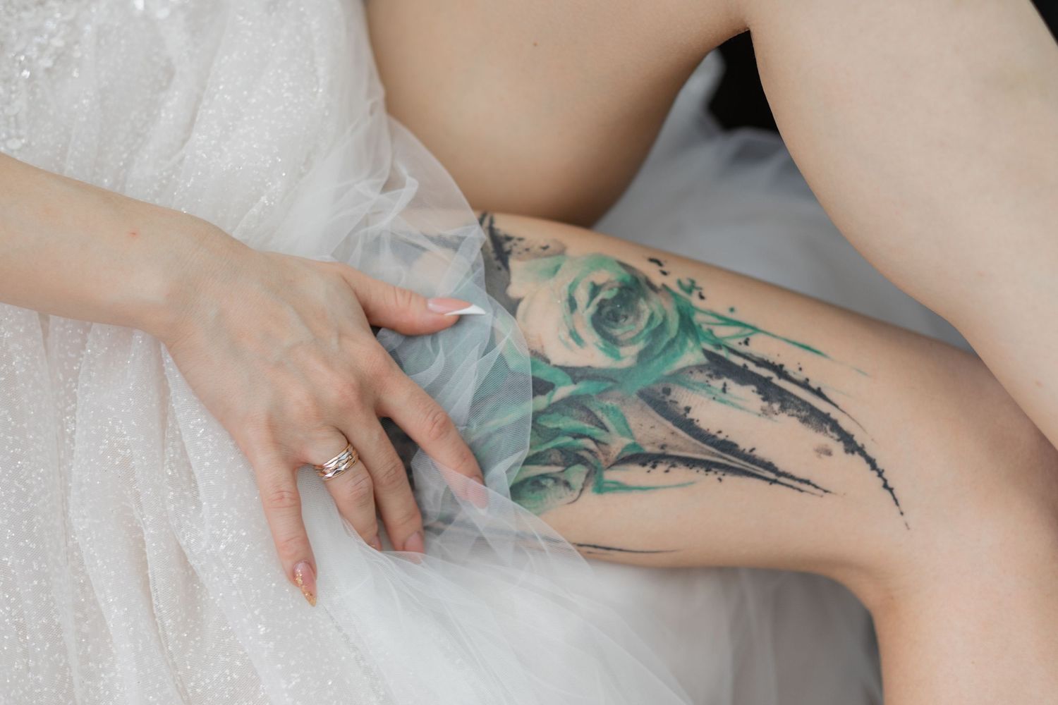 Bride in white dress with watercolor rose tattoo on thigh, wearing diamond ring, elegant wedding detail photo.