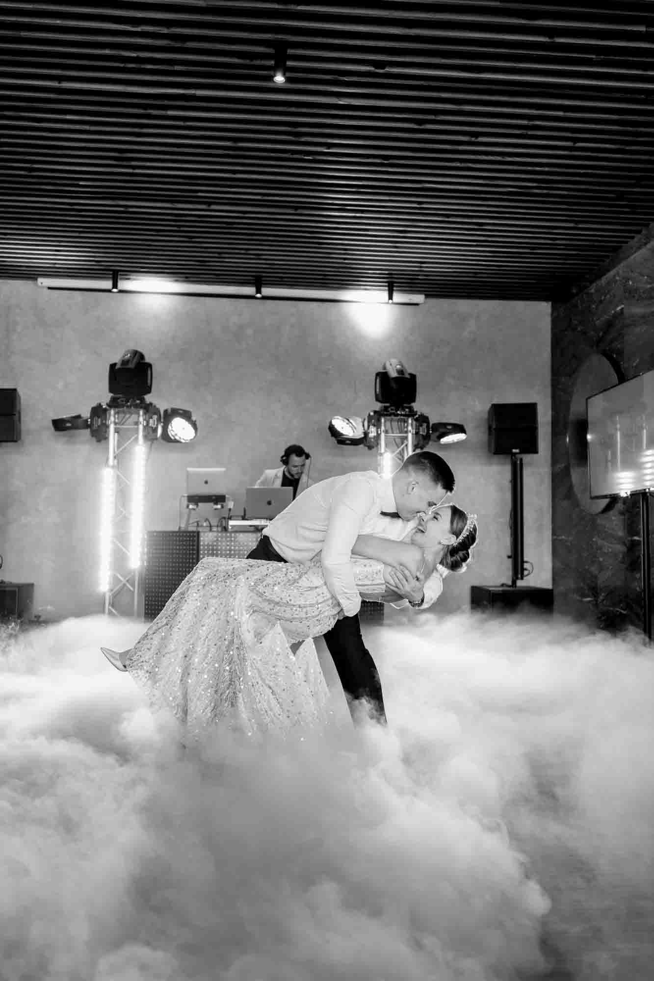 Black and white photo of bride and groom sharing first dance kiss amid dry ice fog with DJ and stage lights behind.