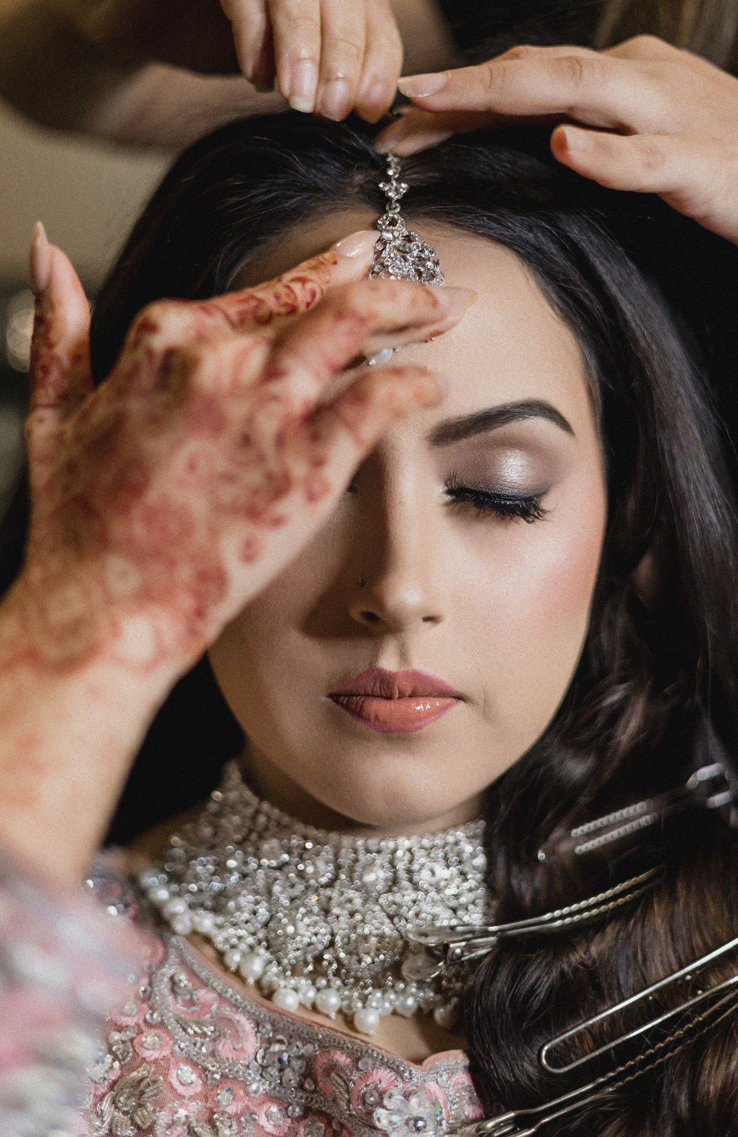 A South Asian bride gets her maang tikka placed as hands with henna apply the jewelry over her elegant bridal makeup.