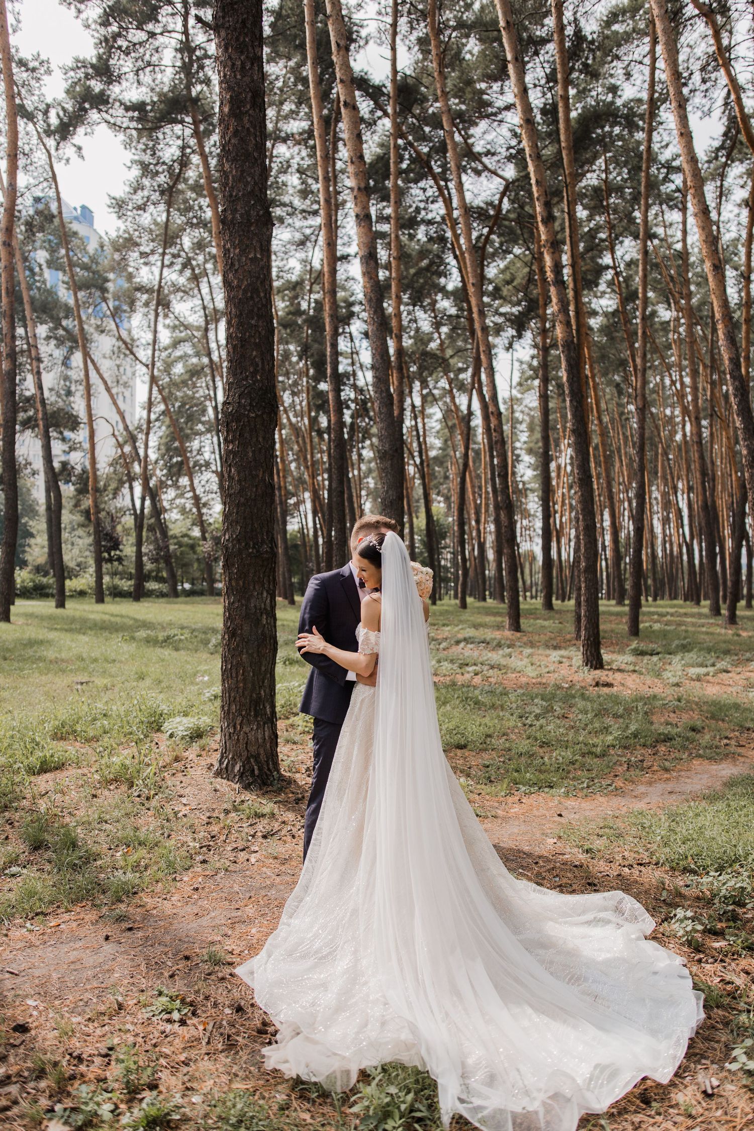 Bride and groom embracing in a pine forest, with her long white veil trailing on the ground.
