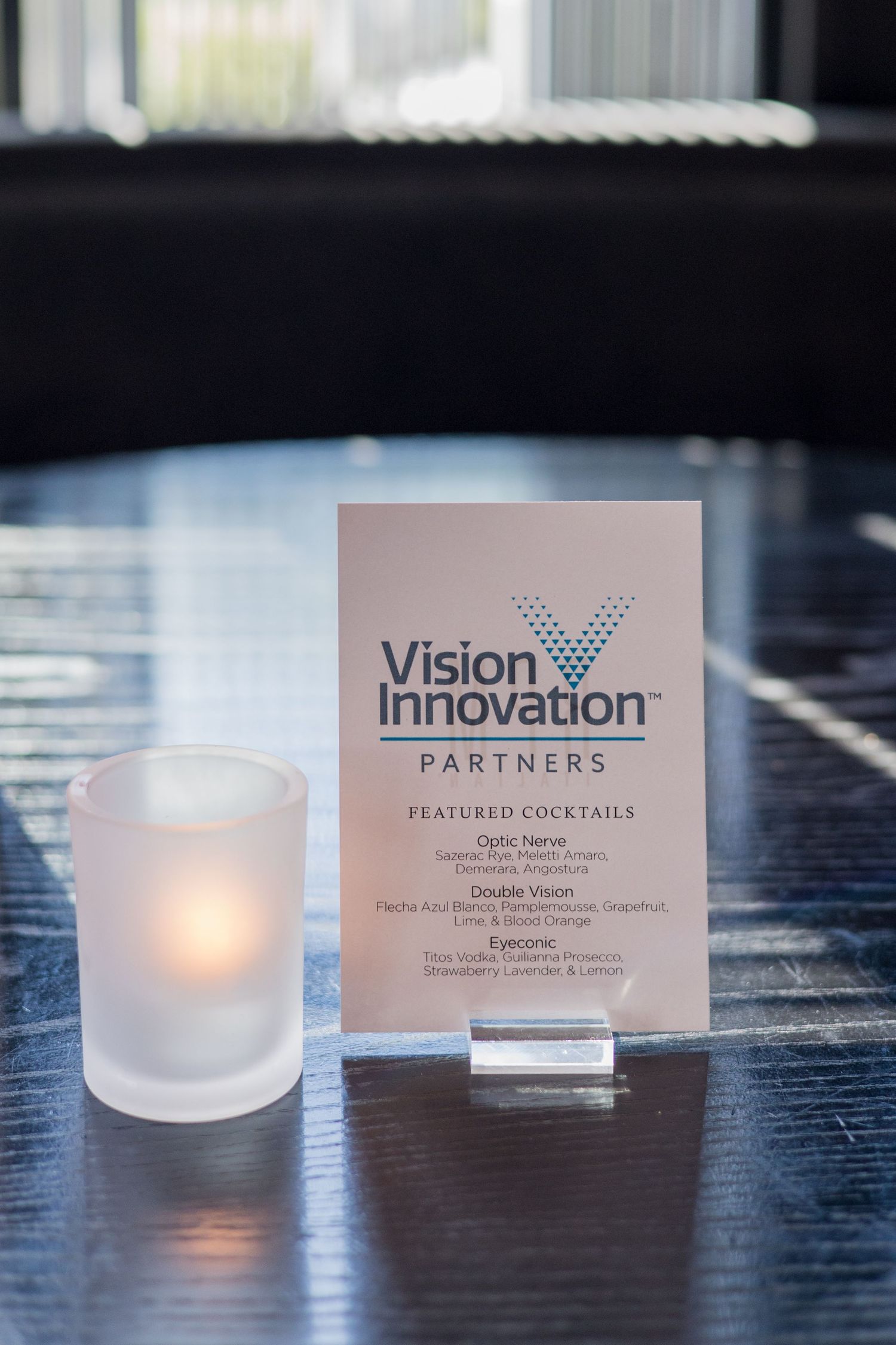 Vision Innovation Partners featured cocktails menu card displayed on a dark table beside a glowing frosted votive candle.