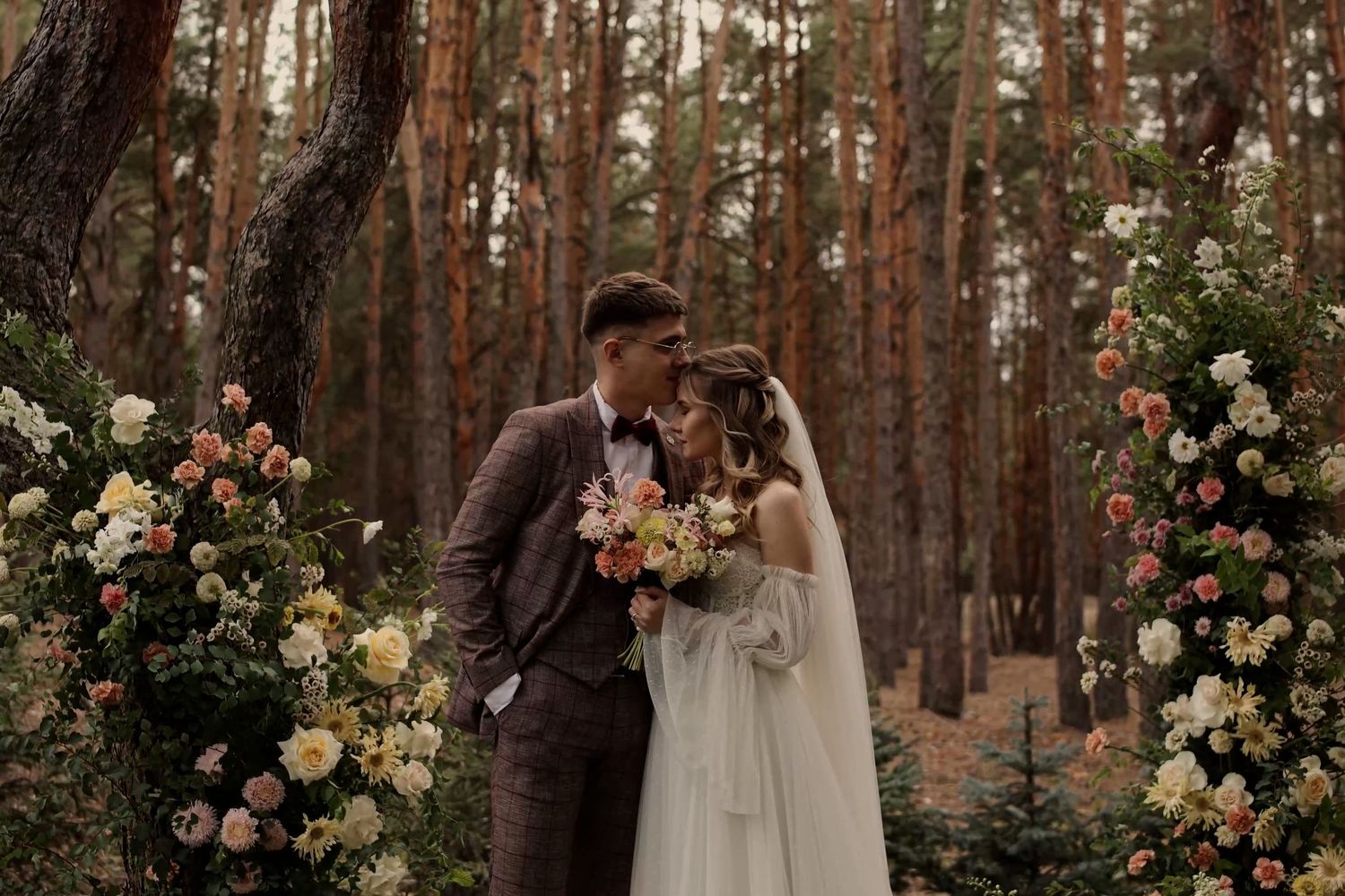 Bride and groom kissing in a forest wedding ceremony surrounded by lush floral arrangements of pink and white roses.