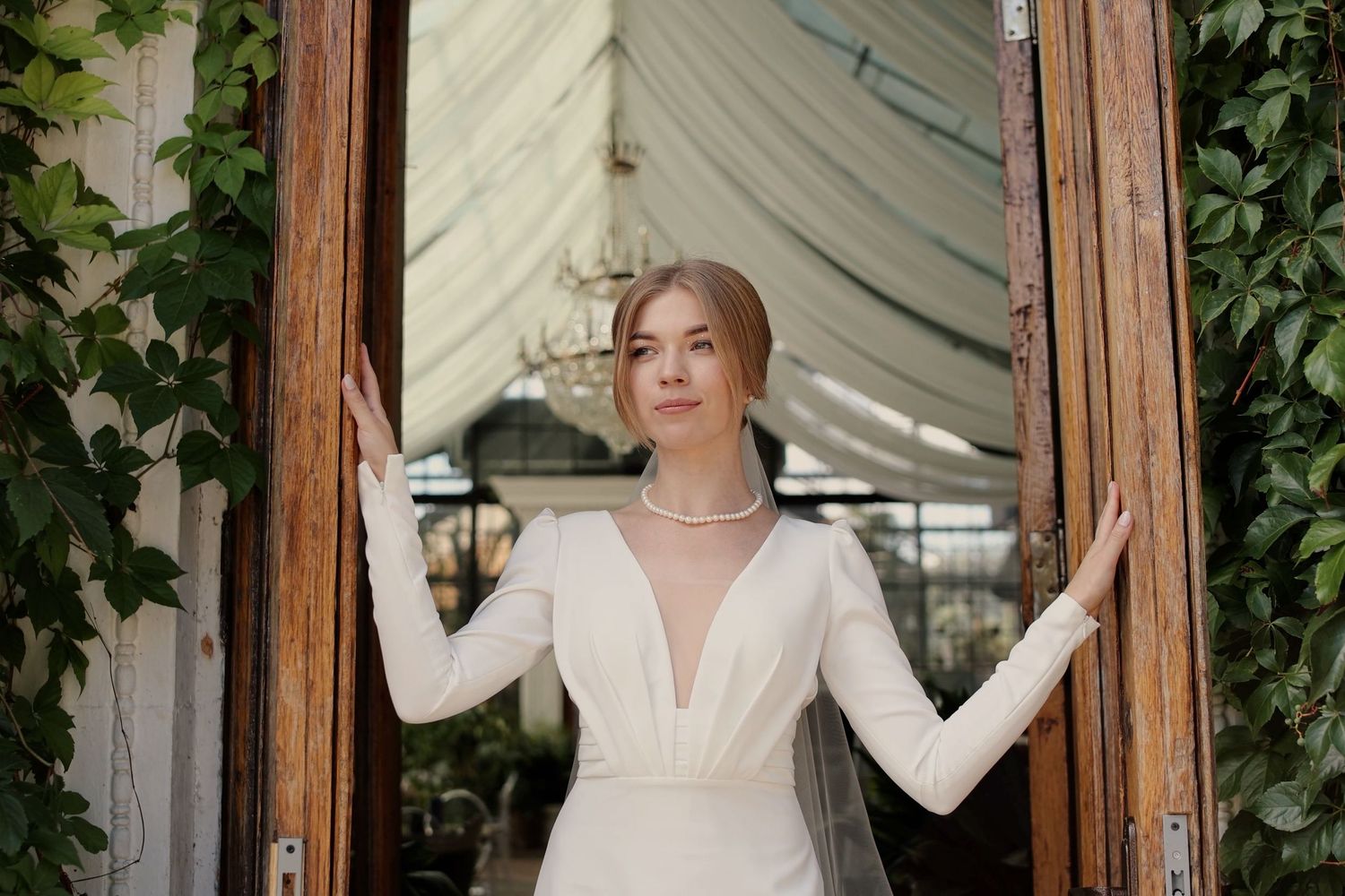 Elegant bride in a white long-sleeve deep V-neck gown with pearl necklace, standing in wooden doorway with green ivy.