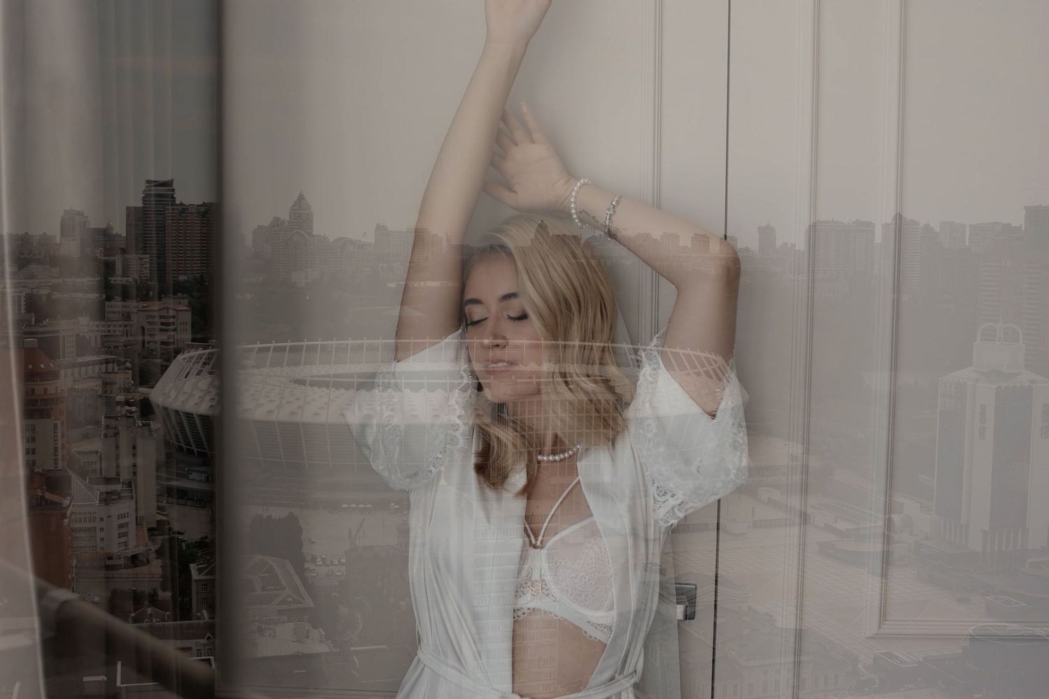 A blonde woman in a white shirt poses gracefully by a window with city views, arms raised above her head.