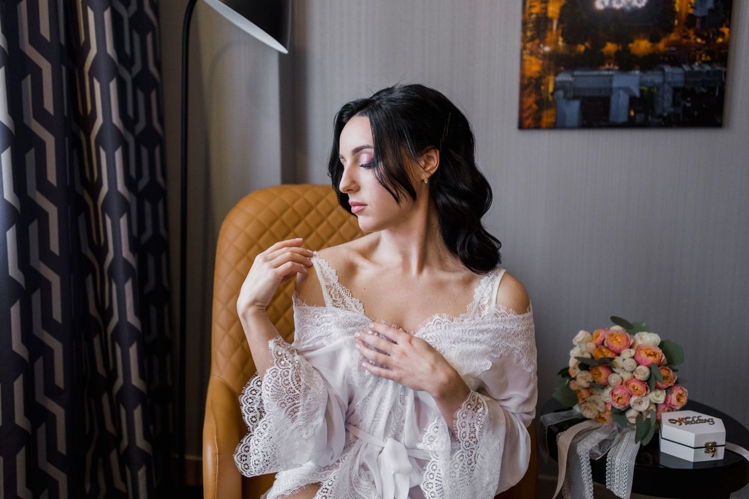 A dark-haired bride in a white lace robe sits in a chair, delicately holding her wedding lingerie near a window.